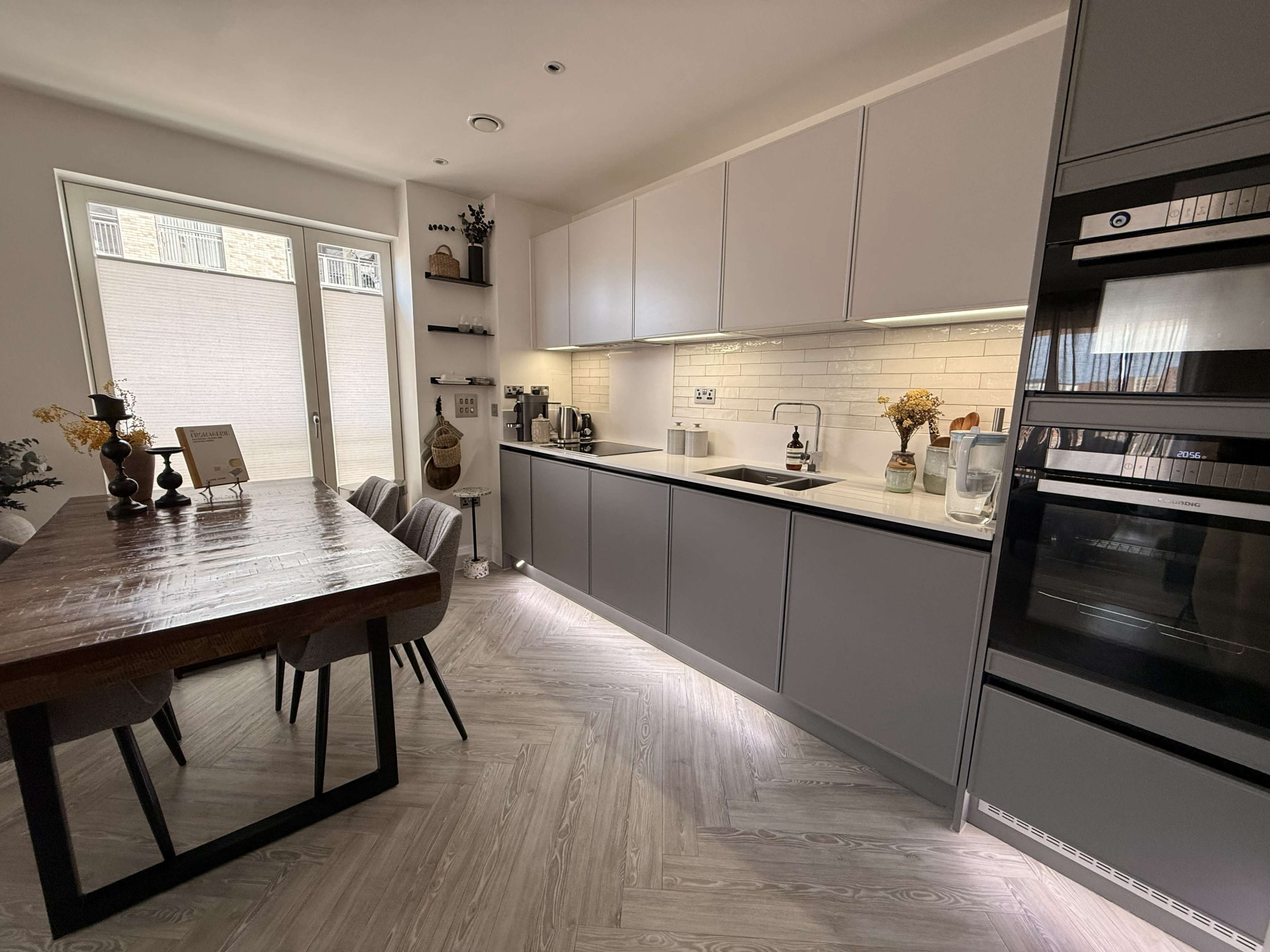 A modern kitchen features a long grey cabinet with built-in appliances, a white countertop, and a wooden dining table with grey chairs.