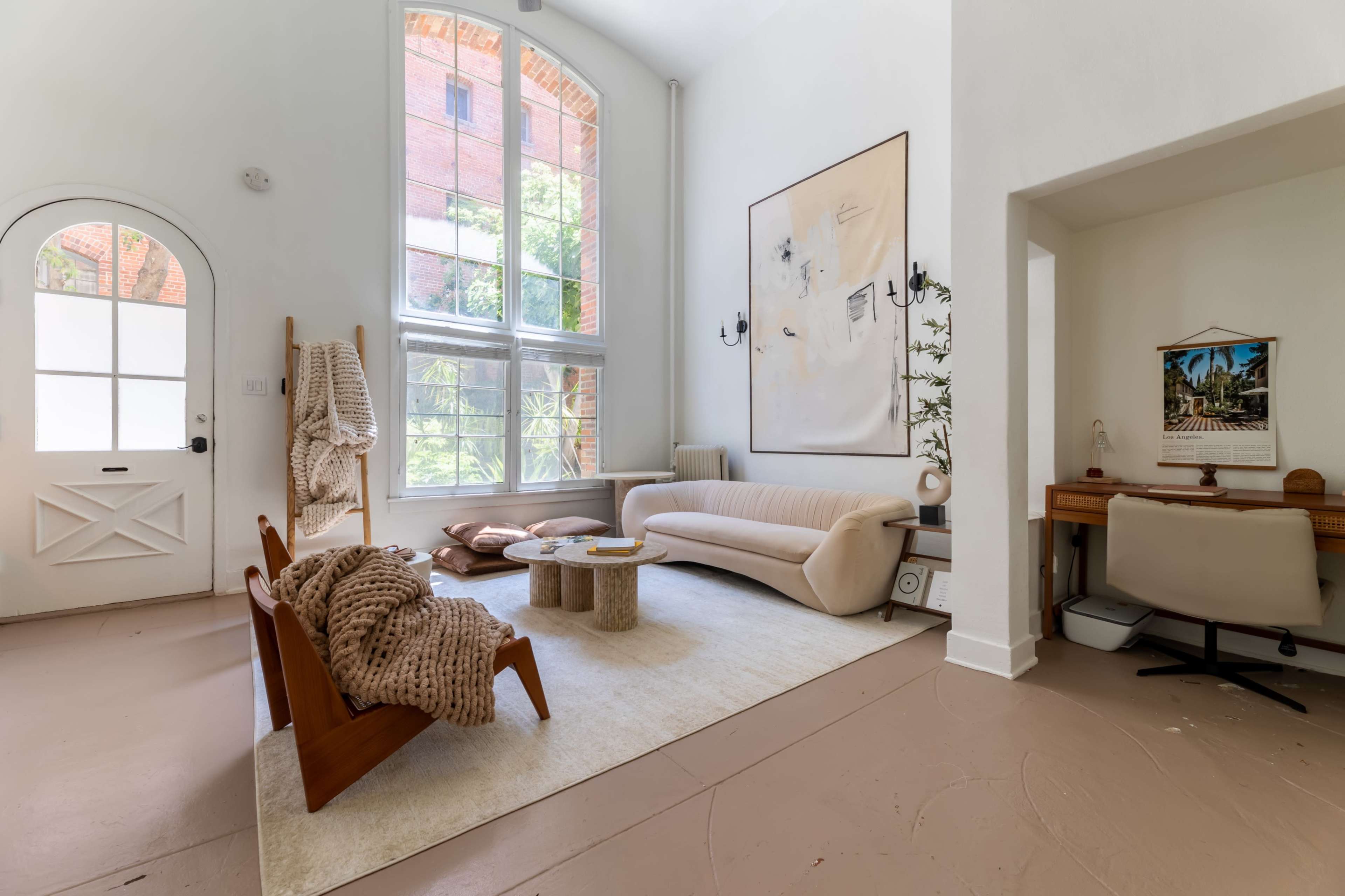 A bright, modern living room featuring a curved sofa, a stone coffee table, and a workspace in the corner, with large windows allowing natural light to fill the space.