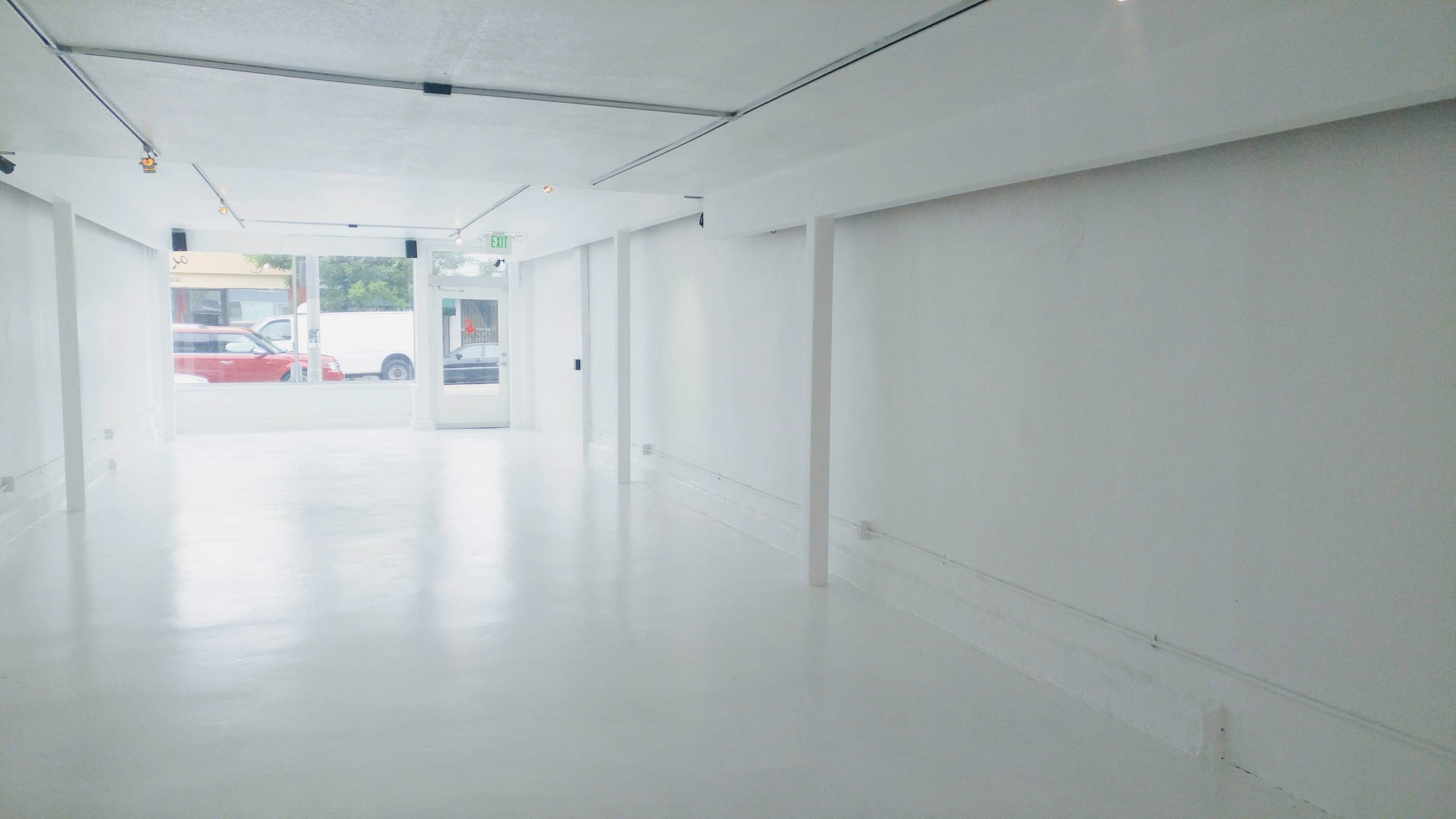 The image shows a large, empty indoor space with white walls and a polished floor, featuring large windows along one side.