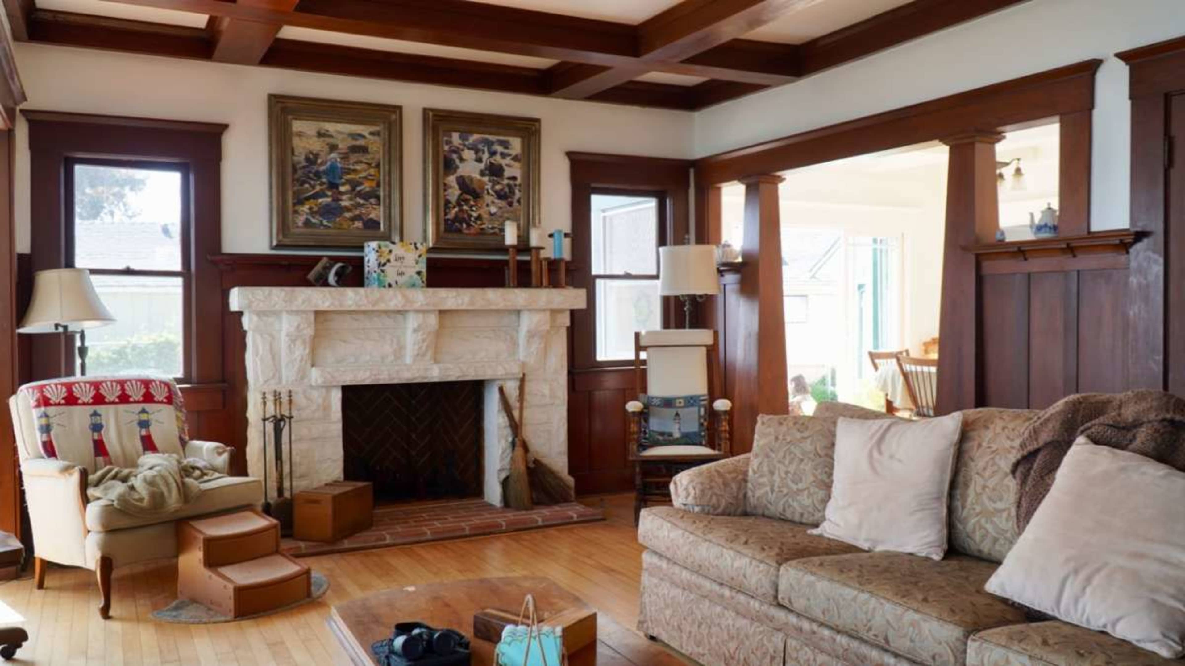 The image shows a cozy living room featuring a stone fireplace, wooden beams on the ceiling, and a comfortable sofa with decorative cushions.
