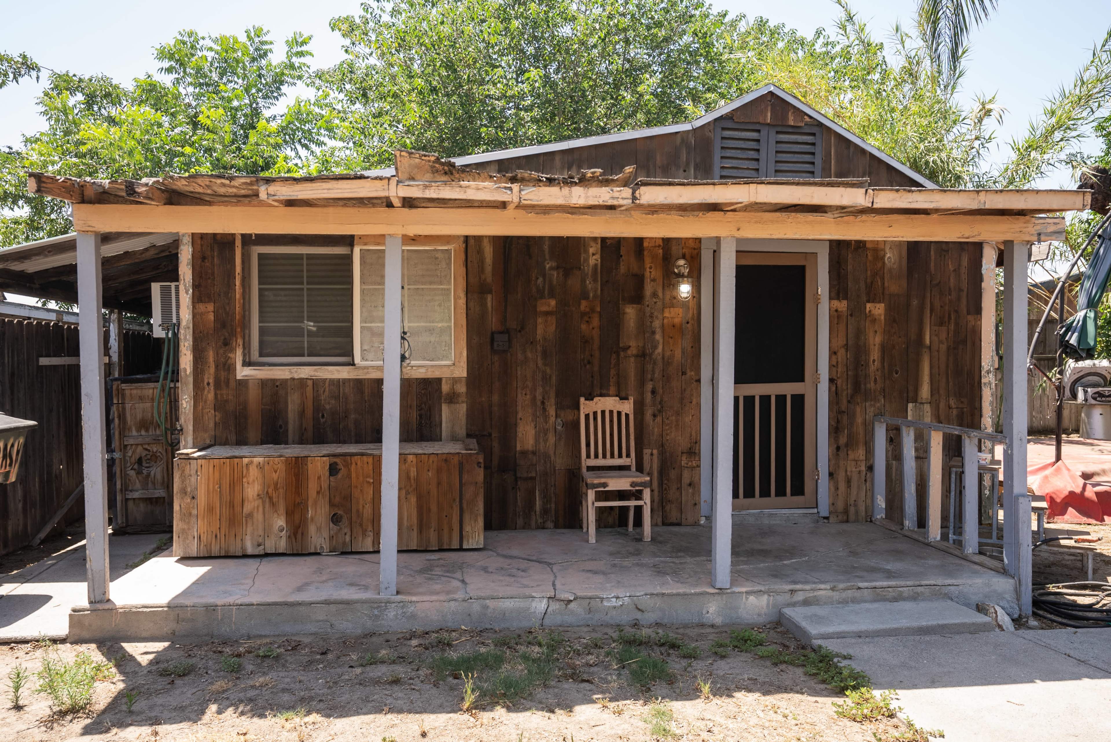 The image shows a rustic wooden cabin with a porch, a single chair, and a front door.