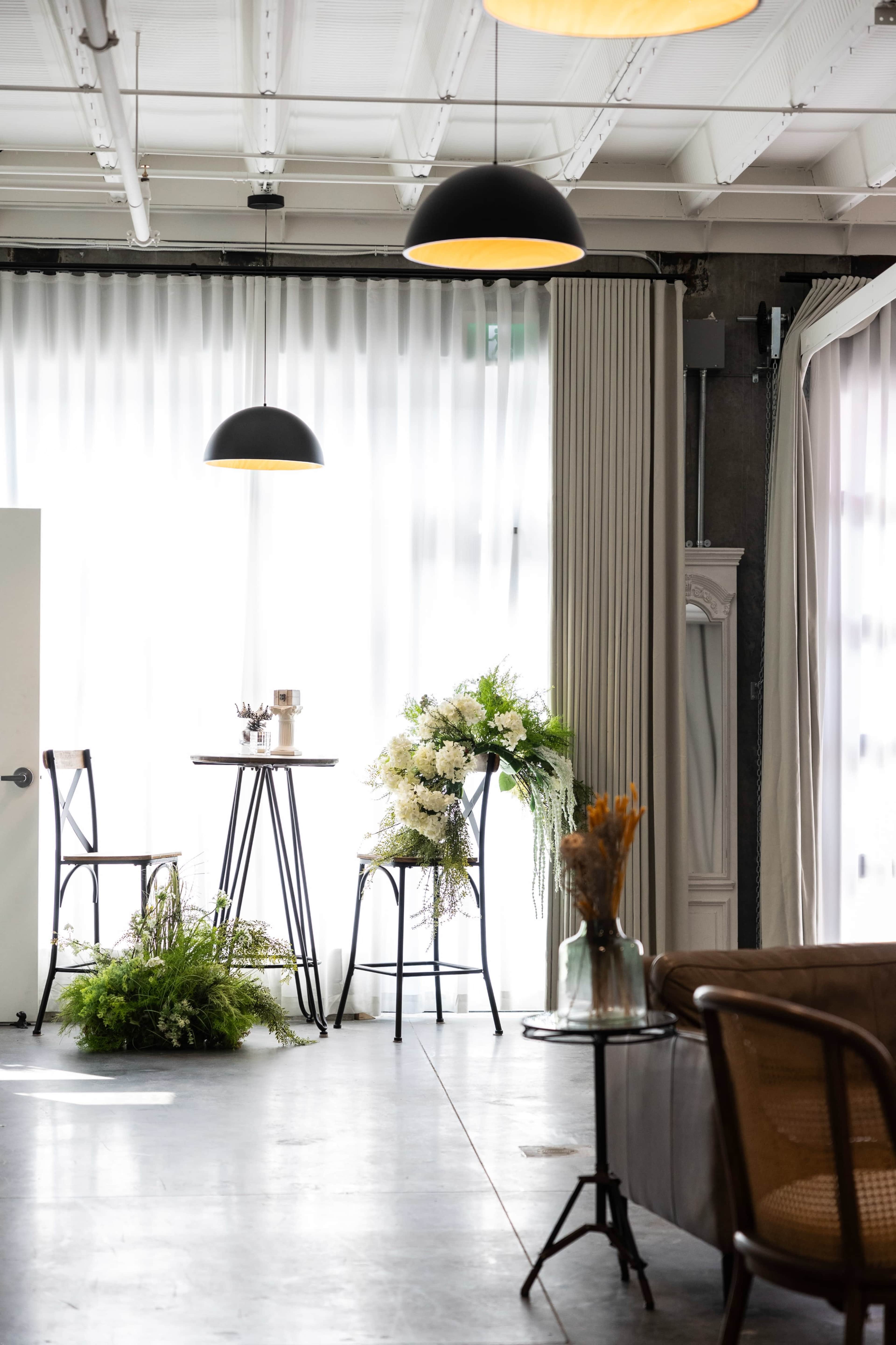 The scene features a modern, well-lit interior with a small table and chairs, adorned with plants and flowers, against a backdrop of large, sheer curtains.