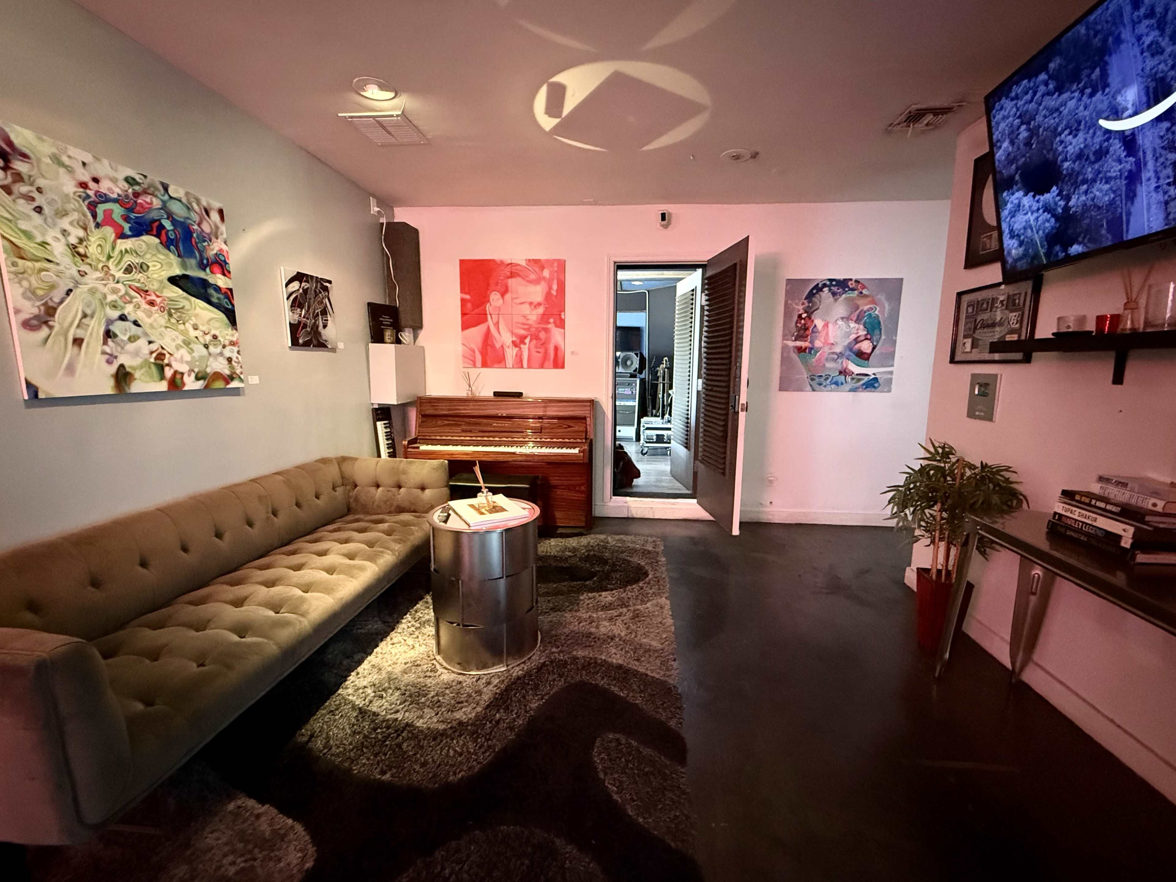 The image shows a modern lounge area with a tufted sofa, a round table, a piano, colorful artwork on the walls, and a doorway leading to another room.