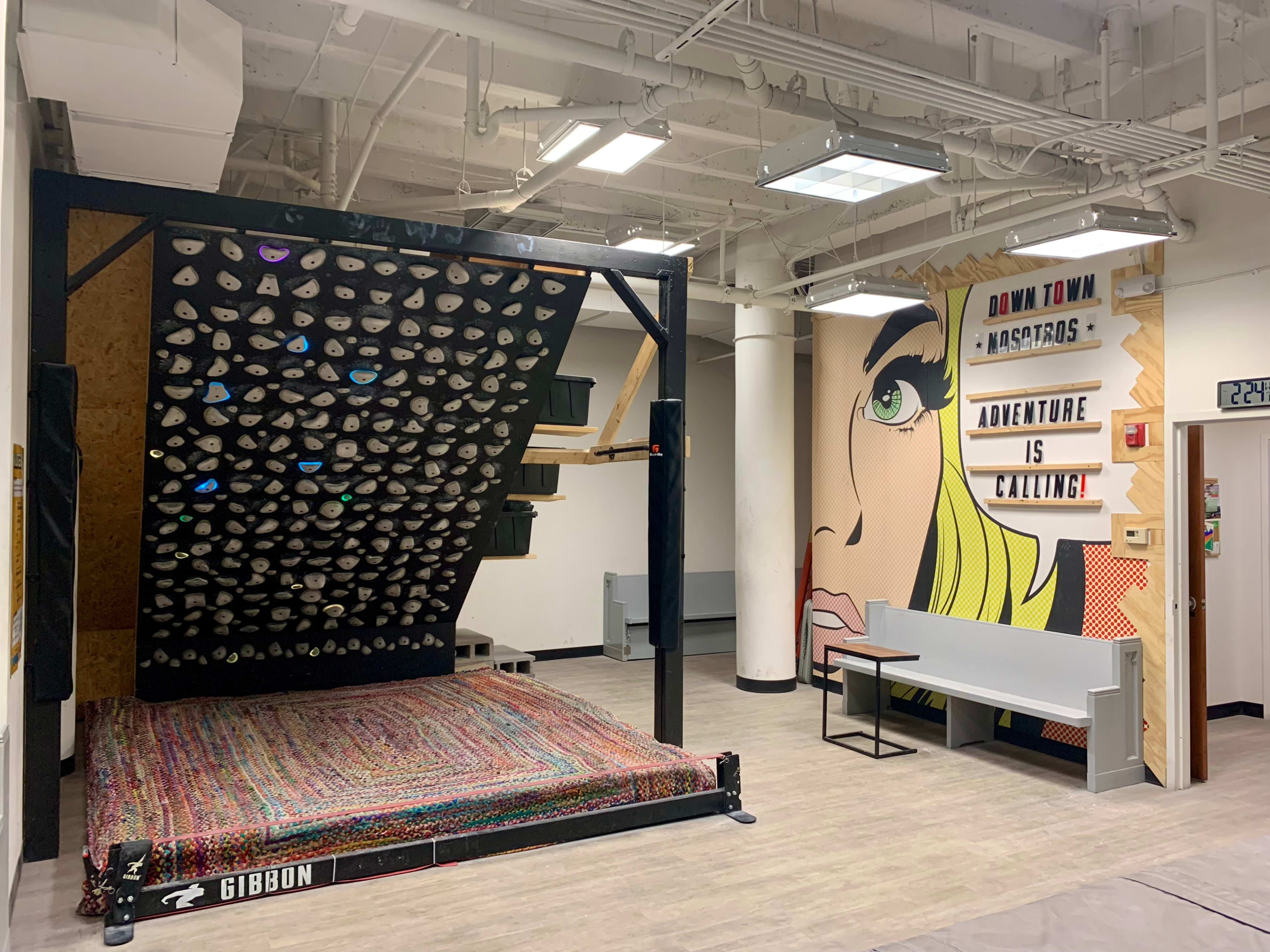 The image shows an indoor rock climbing wall with a colorful textured mat beneath it, alongside a mural featuring a stylized comic book character and motivational text.