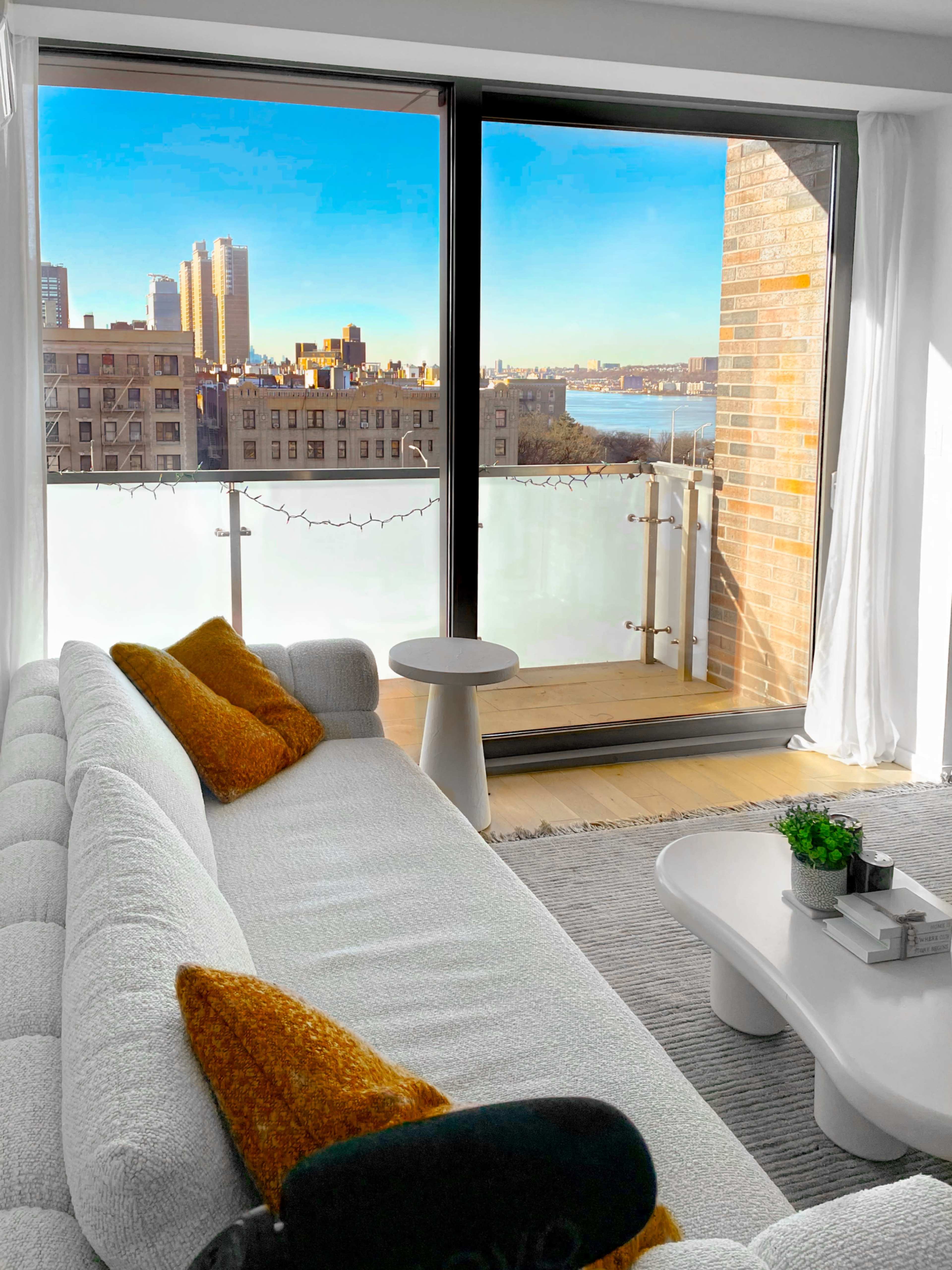 A modern living room features a light-colored sofa with orange accents, overlooking a cityscape through large glass windows.