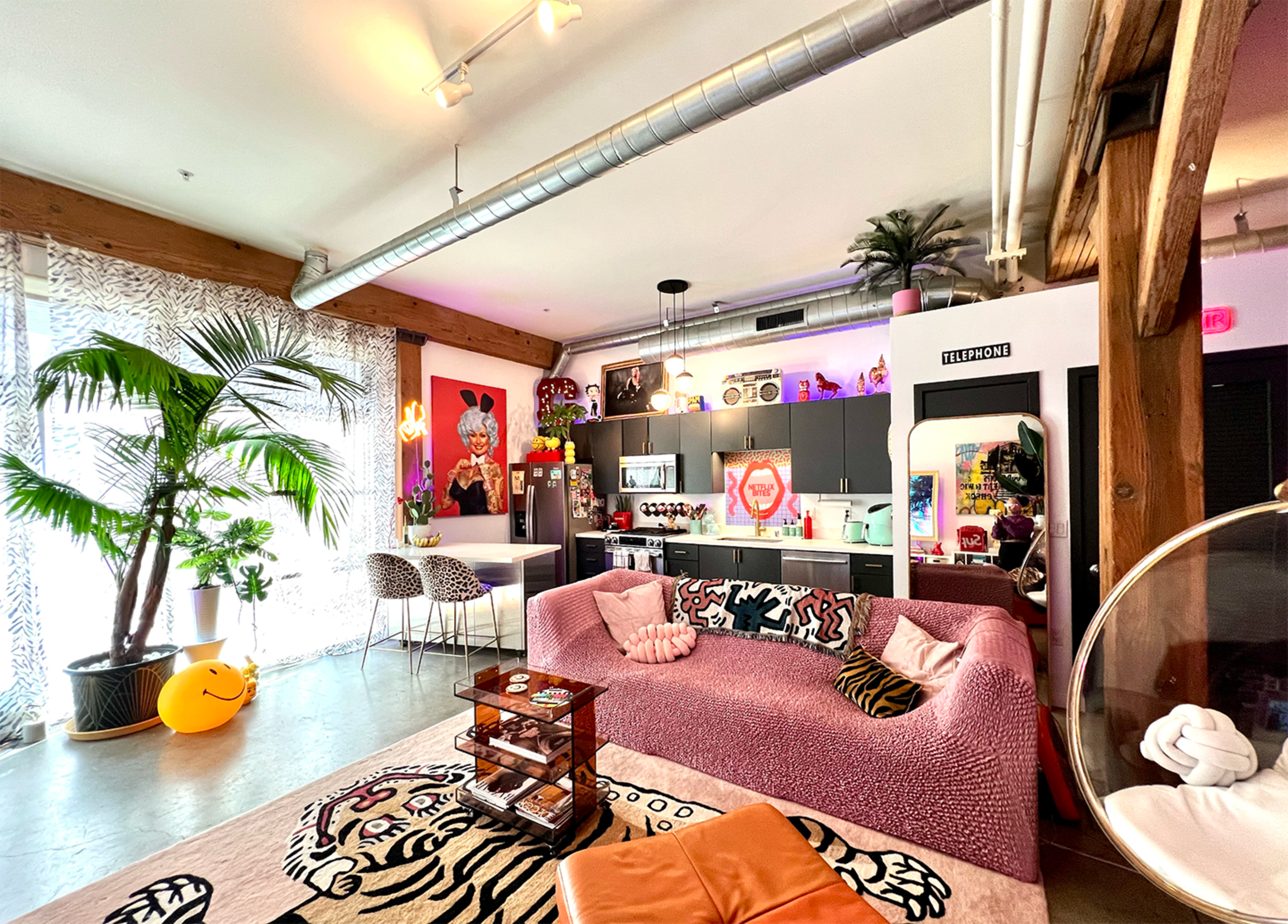 The image shows a vibrant, eclectic living space featuring a pink sofa, decorative plants, and a mixture of modern and retro decor elements.