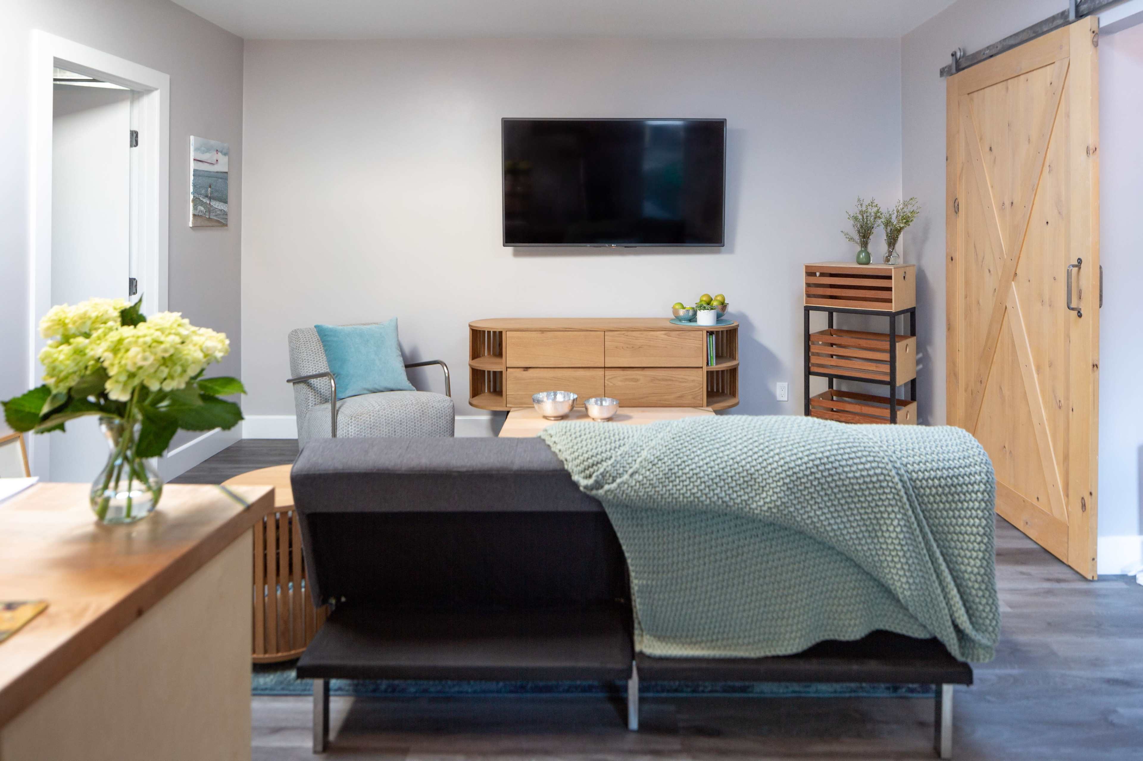 A modern living room features a wall-mounted TV, a wooden media console, a gray sofa with a knitted throw, and natural light coming from a door leading outside.