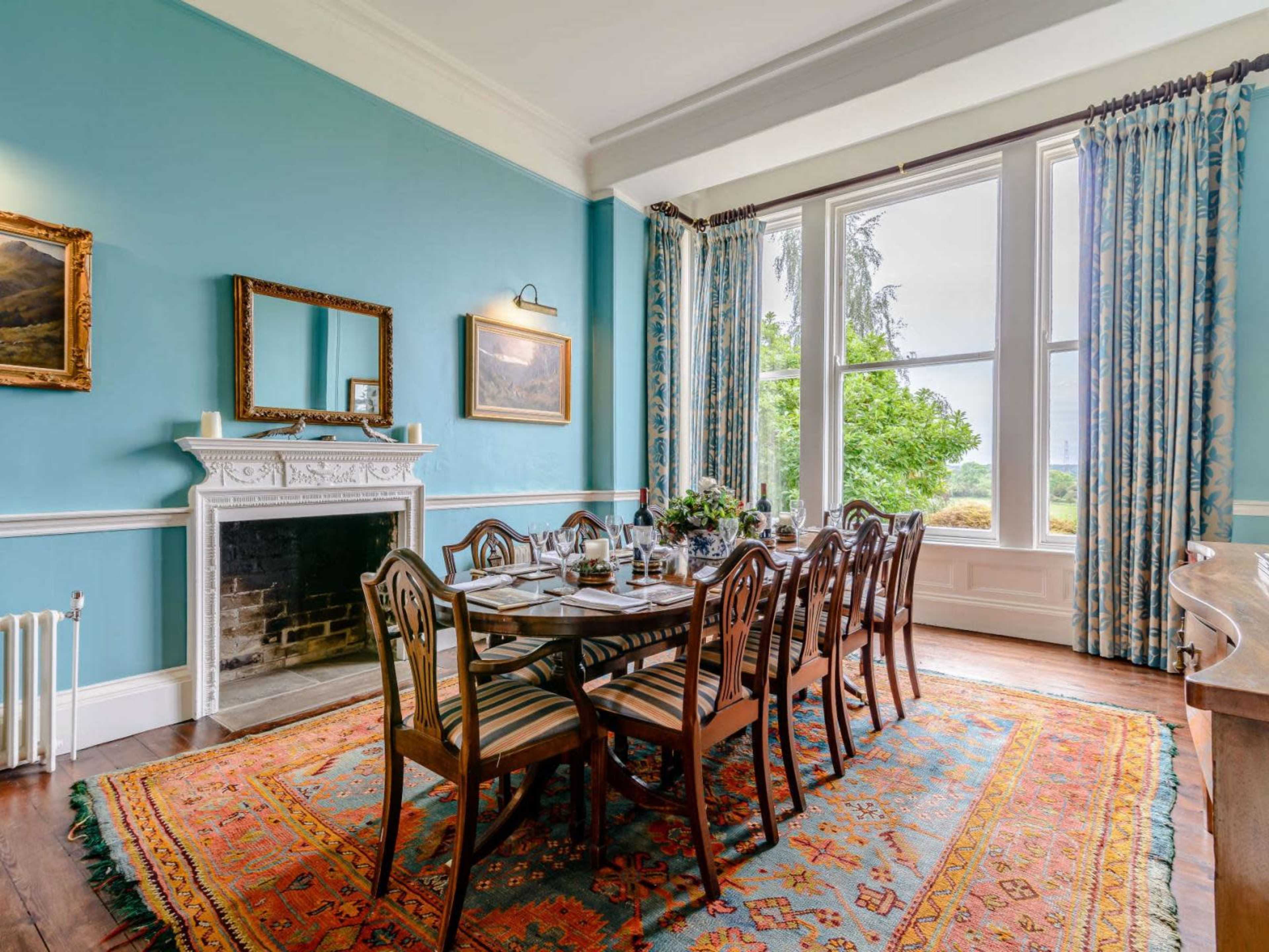 The dining room features a long table surrounded by wooden chairs, a large window with scenic views, and a decorative area rug on the hardwood floor.