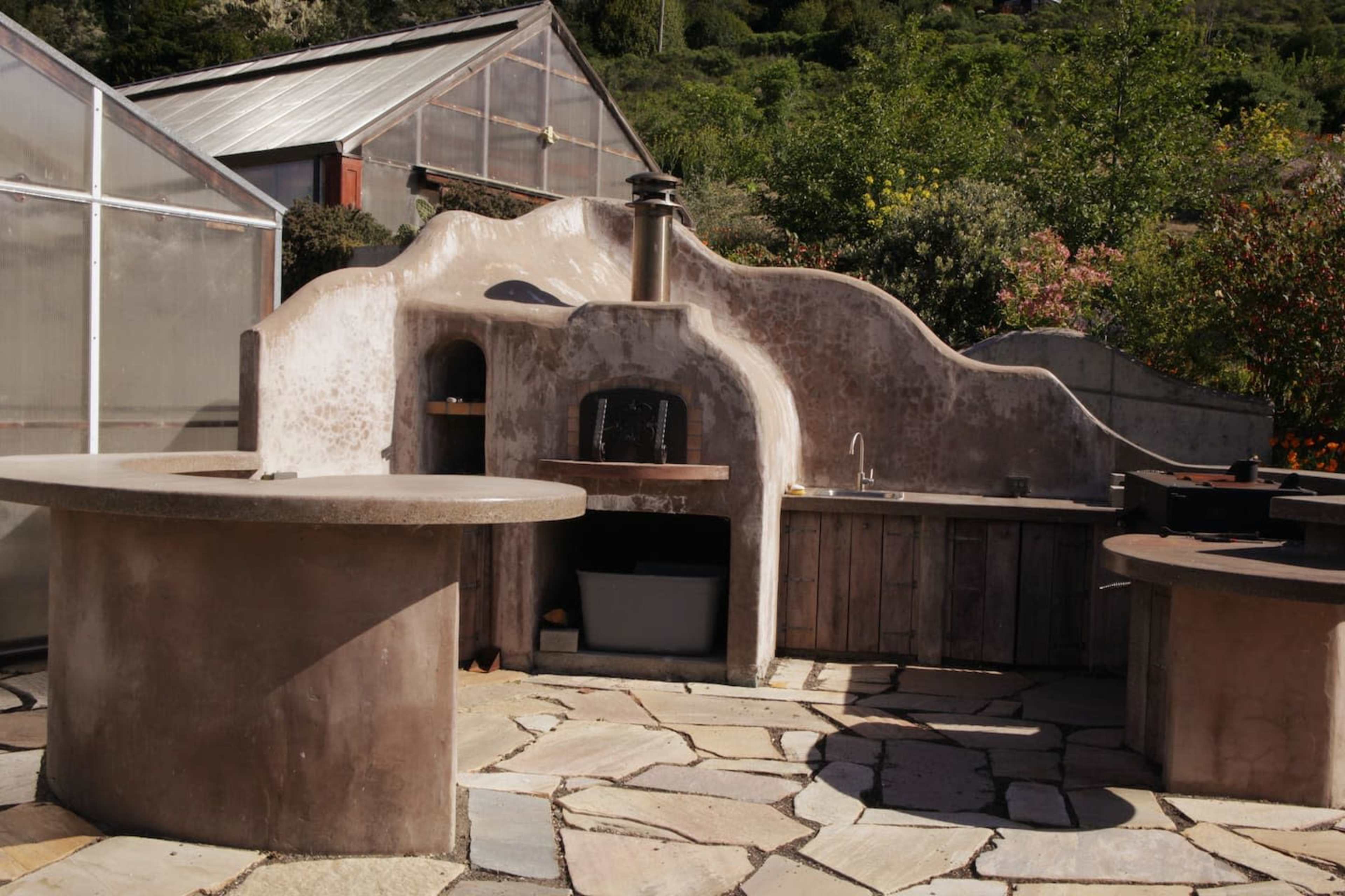 An outdoor kitchen area with circular stone tables, a central cooking station, and a sculptural wall behind it, all set on a flagstone patio surrounded by greenery.