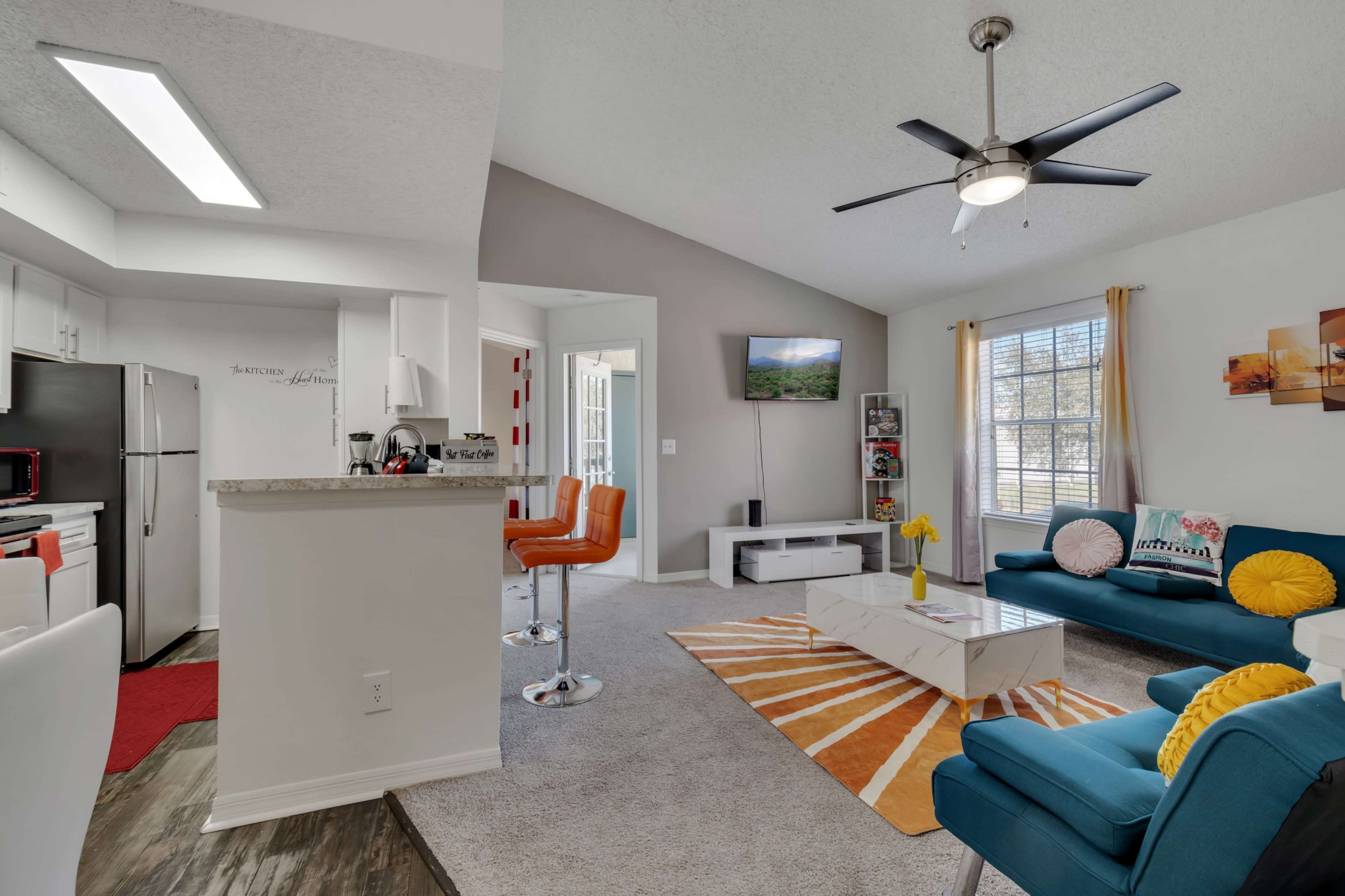 A modern living room with a blue sofa, orange accent chairs, a white coffee table on a sunburst rug, and a well-equipped kitchen visible in the background.