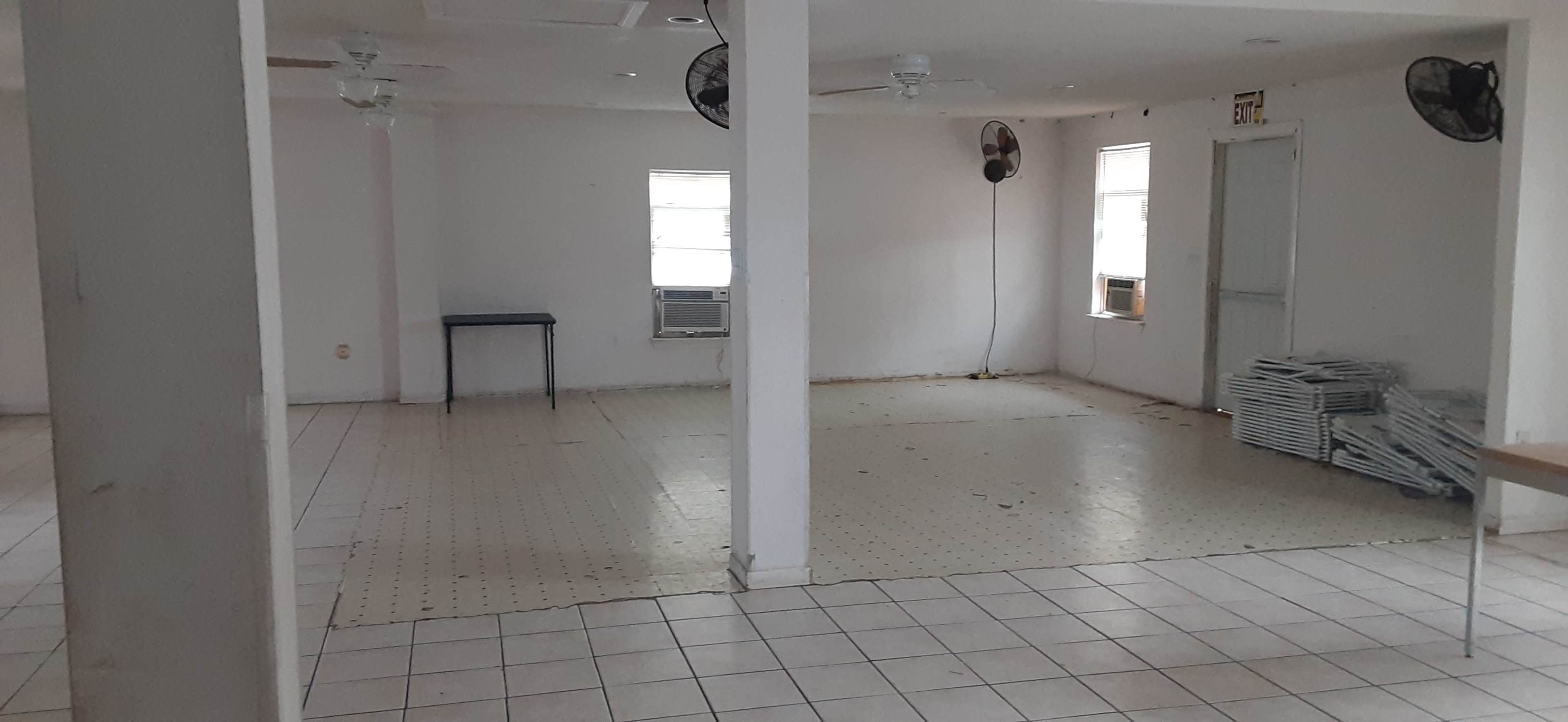 The image shows an empty room with tiled floors, white walls, and two ceiling fans, featuring a table in the corner and an air conditioning unit.
