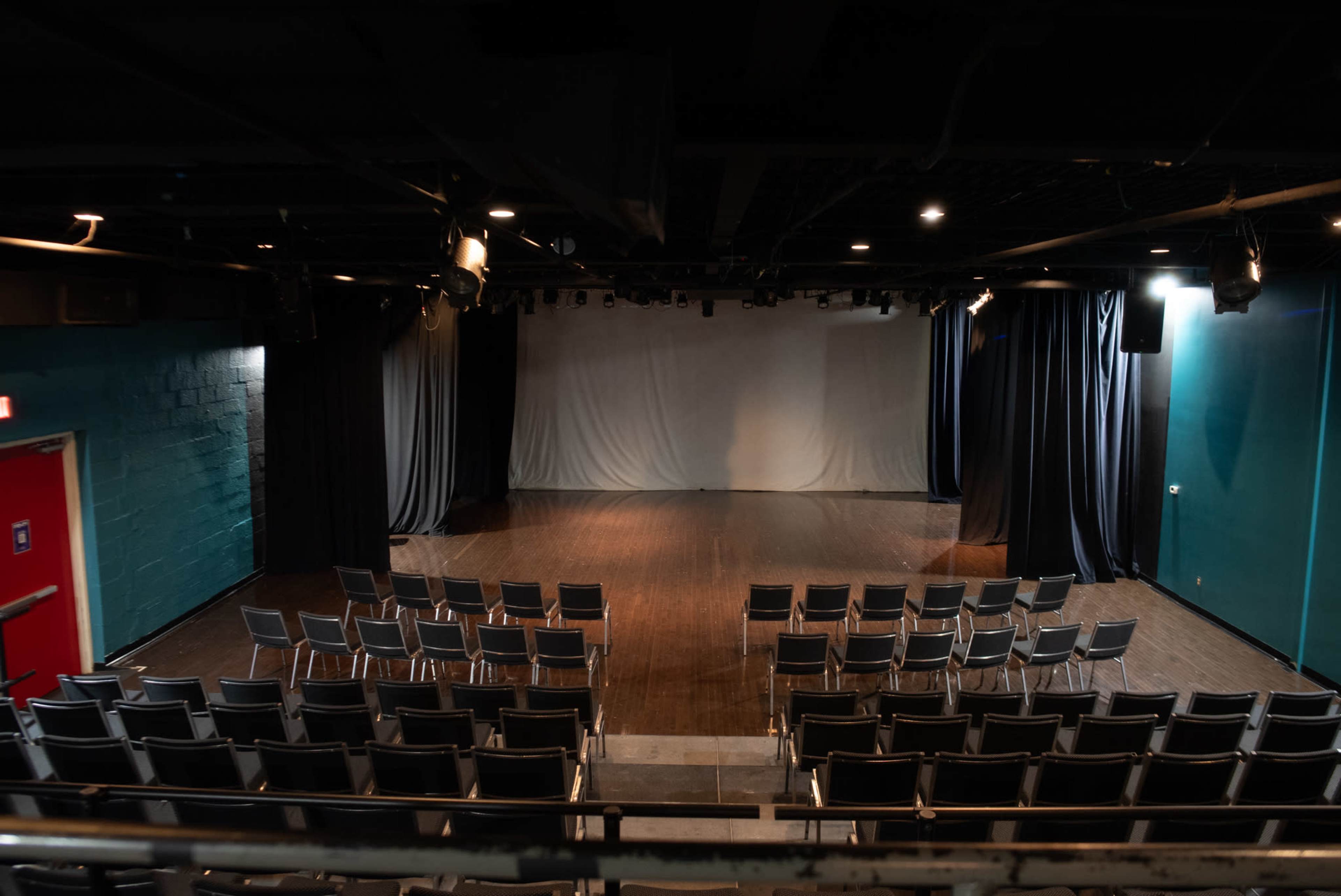 An empty theater stage with rows of metal chairs facing a blank backdrop.