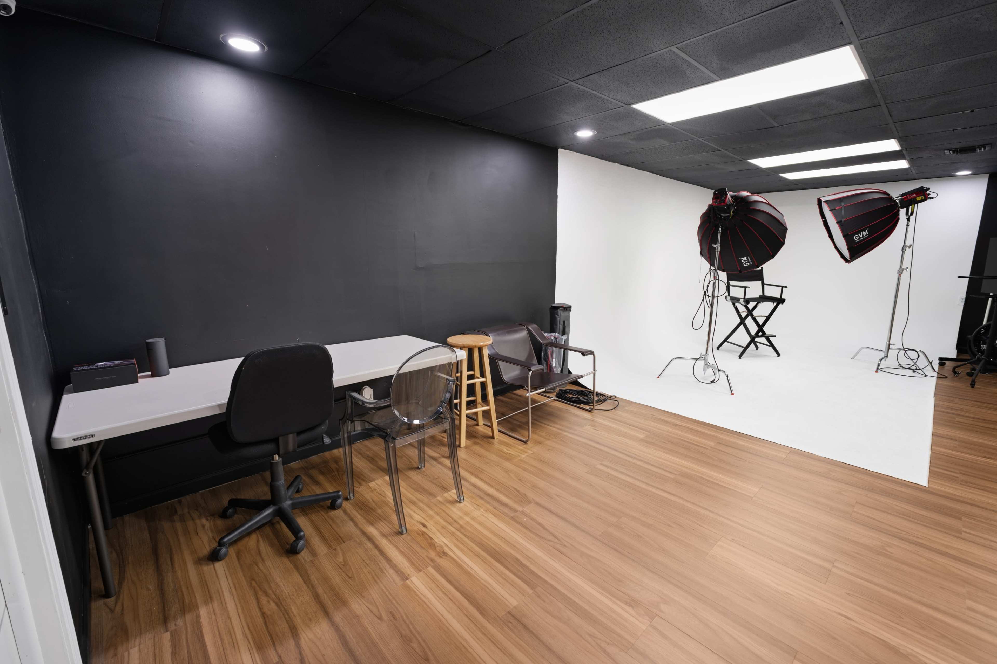 The image shows a photography studio with a black wall, a white backdrop, and two lighting setups, alongside a desk, chair, and stool.
