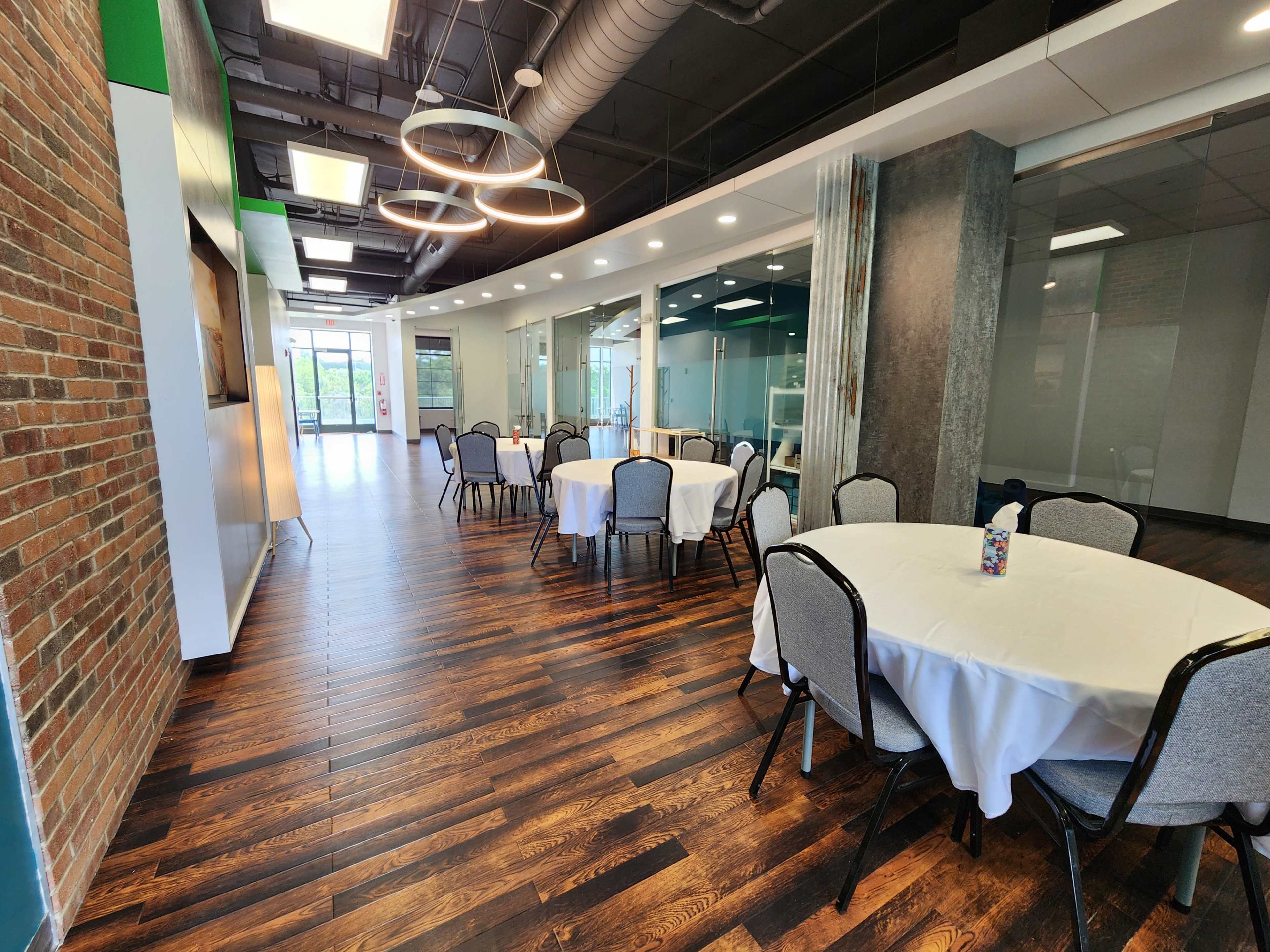 A modern meeting room with round tables covered in white tablecloths, hardwood floors, and large windows allowing natural light.