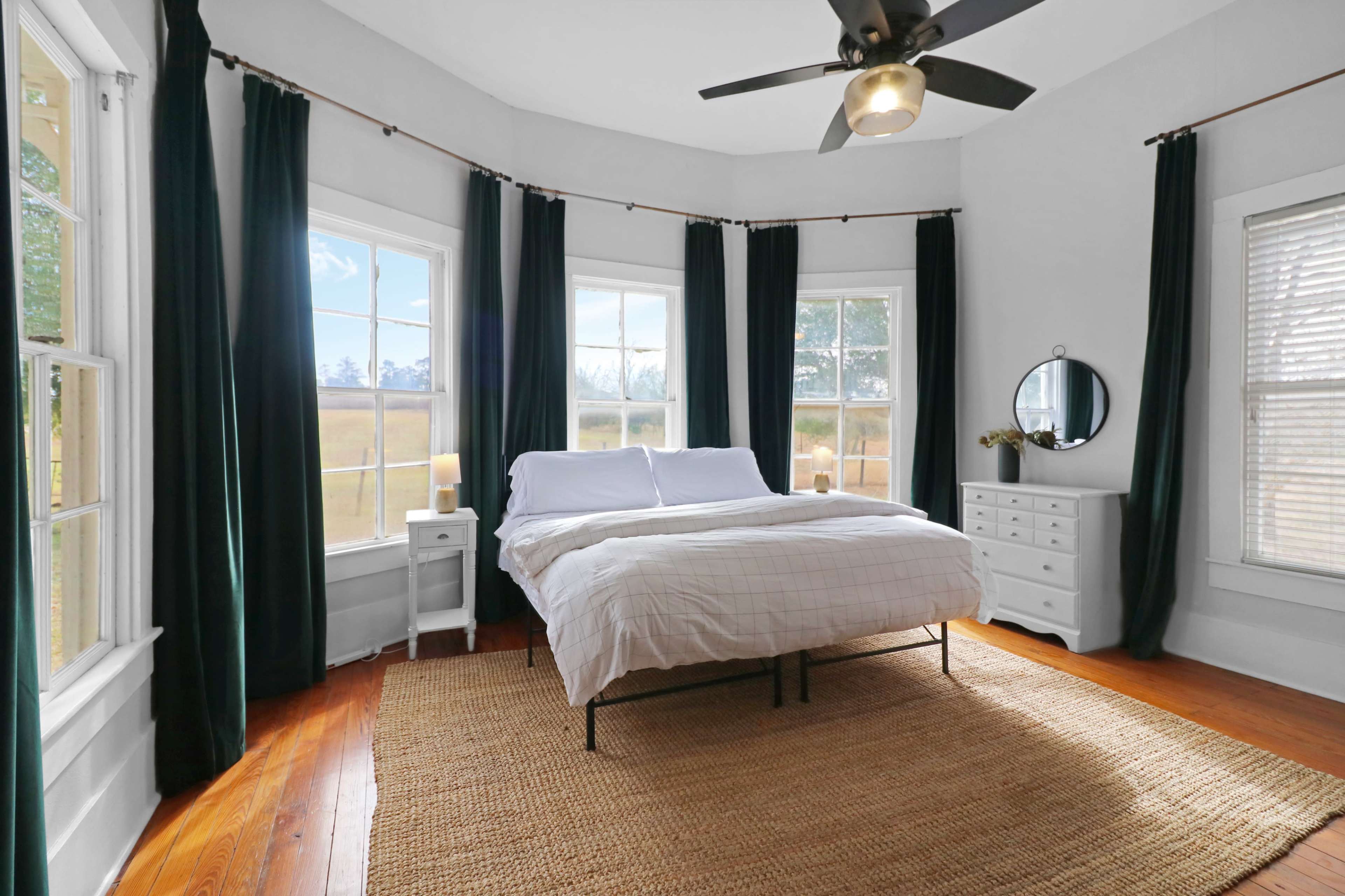 A bedroom features a bed with a white duvet, surrounded by large windows, curtains, a dresser, and a mirror.