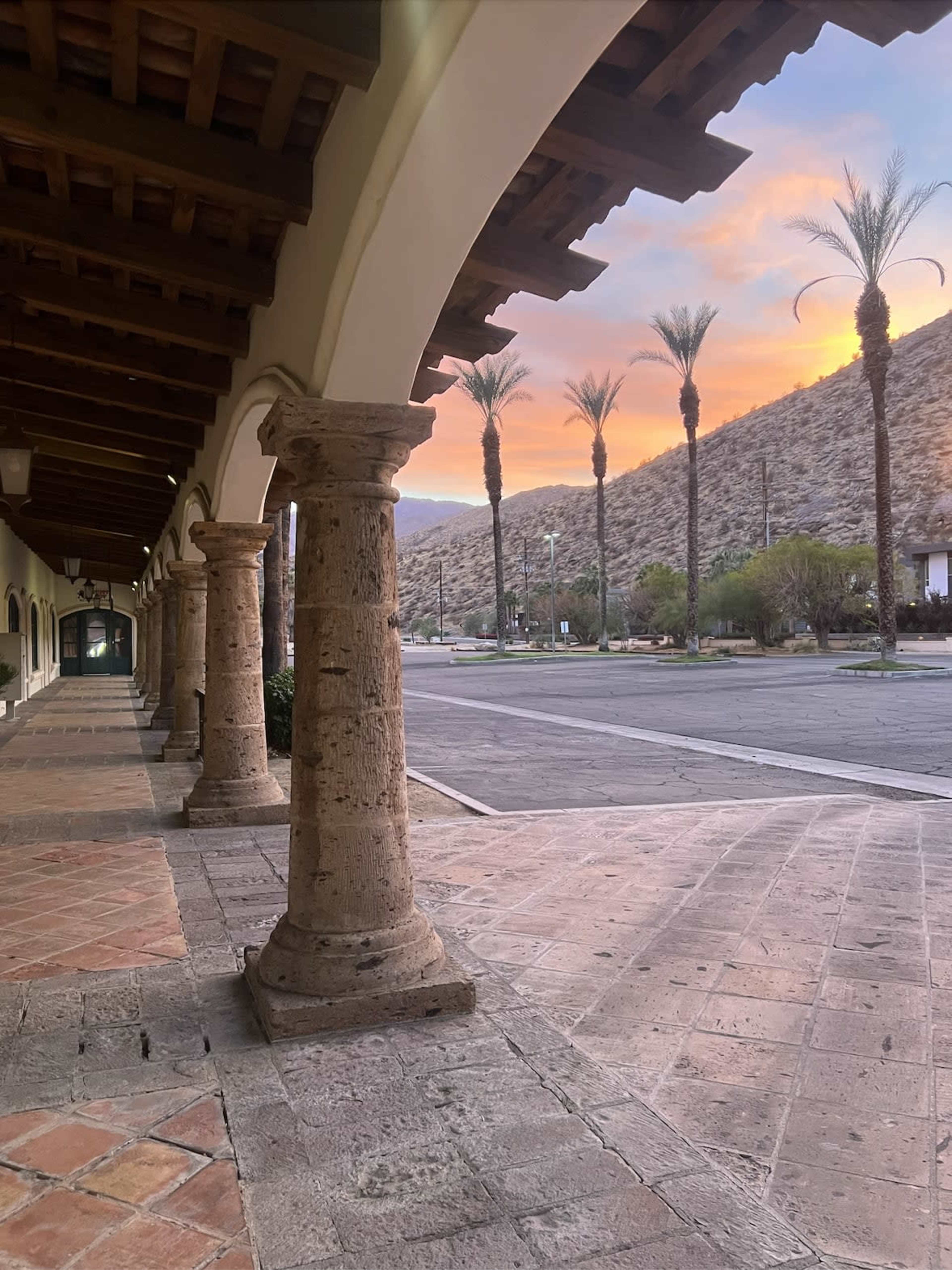 The image shows a stone-paved walkway lined with columns, leading to a street framed by palm trees and a mountainous backdrop under a colorful sky at sunset.