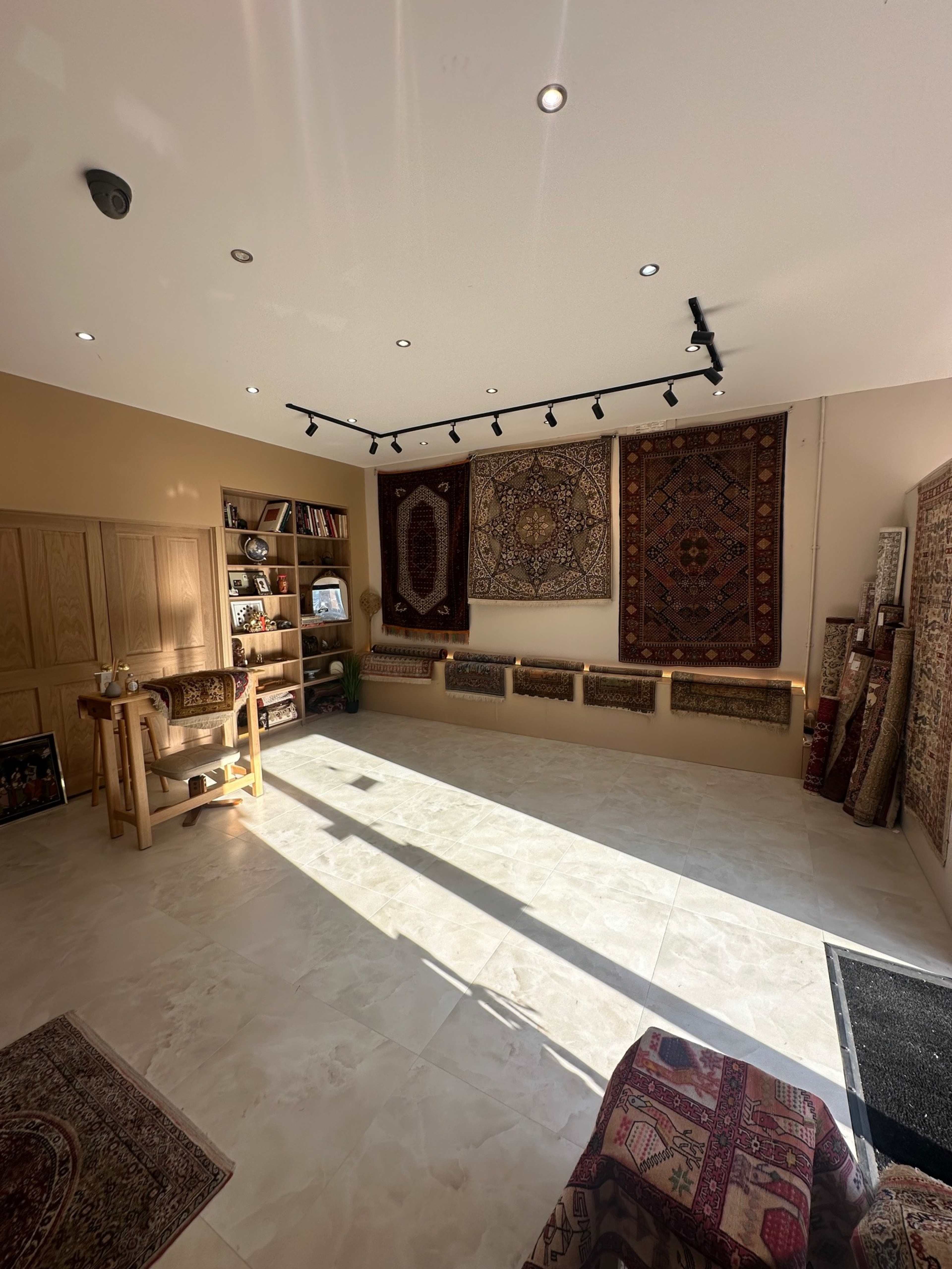 The image shows a spacious, well-lit room featuring decorative rugs hung on the walls and a seating area with a wooden table and shelves.