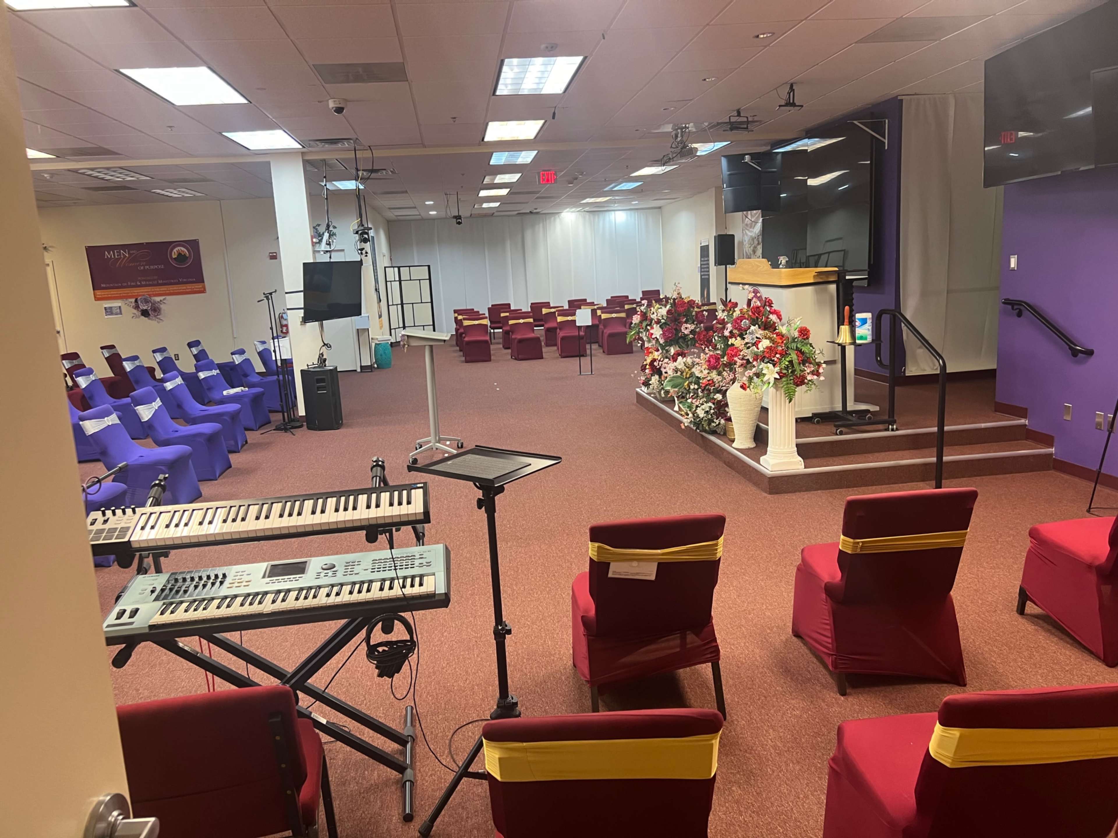 The image shows a spacious room set up for a gathering, featuring rows of purple and red chairs, a keyboard, and a floral arrangement near a podium.