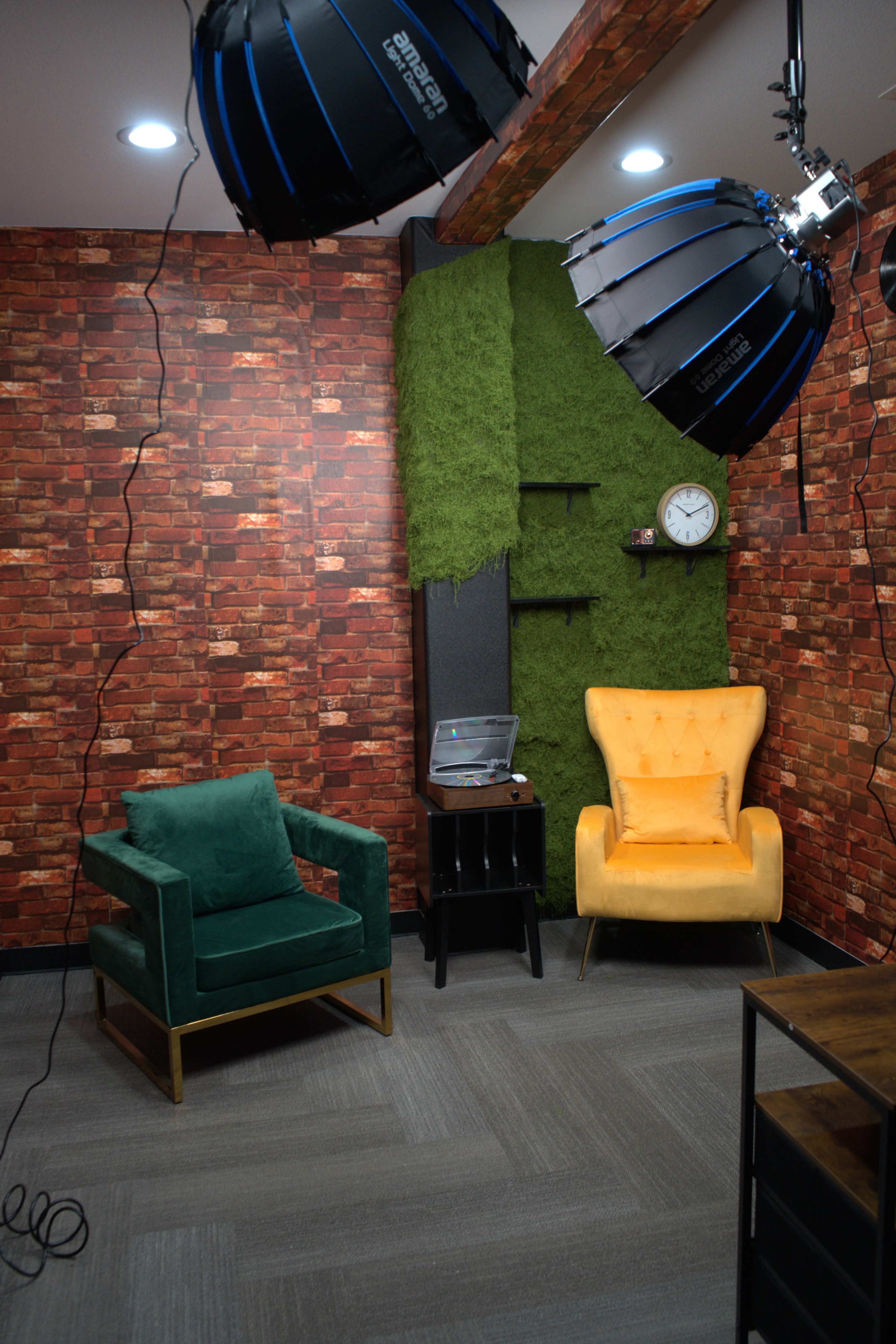 The image shows a well-lit studio space featuring two chairs—one green and one yellow—against a brick-patterned wall with a patch of greenery and a wall clock.