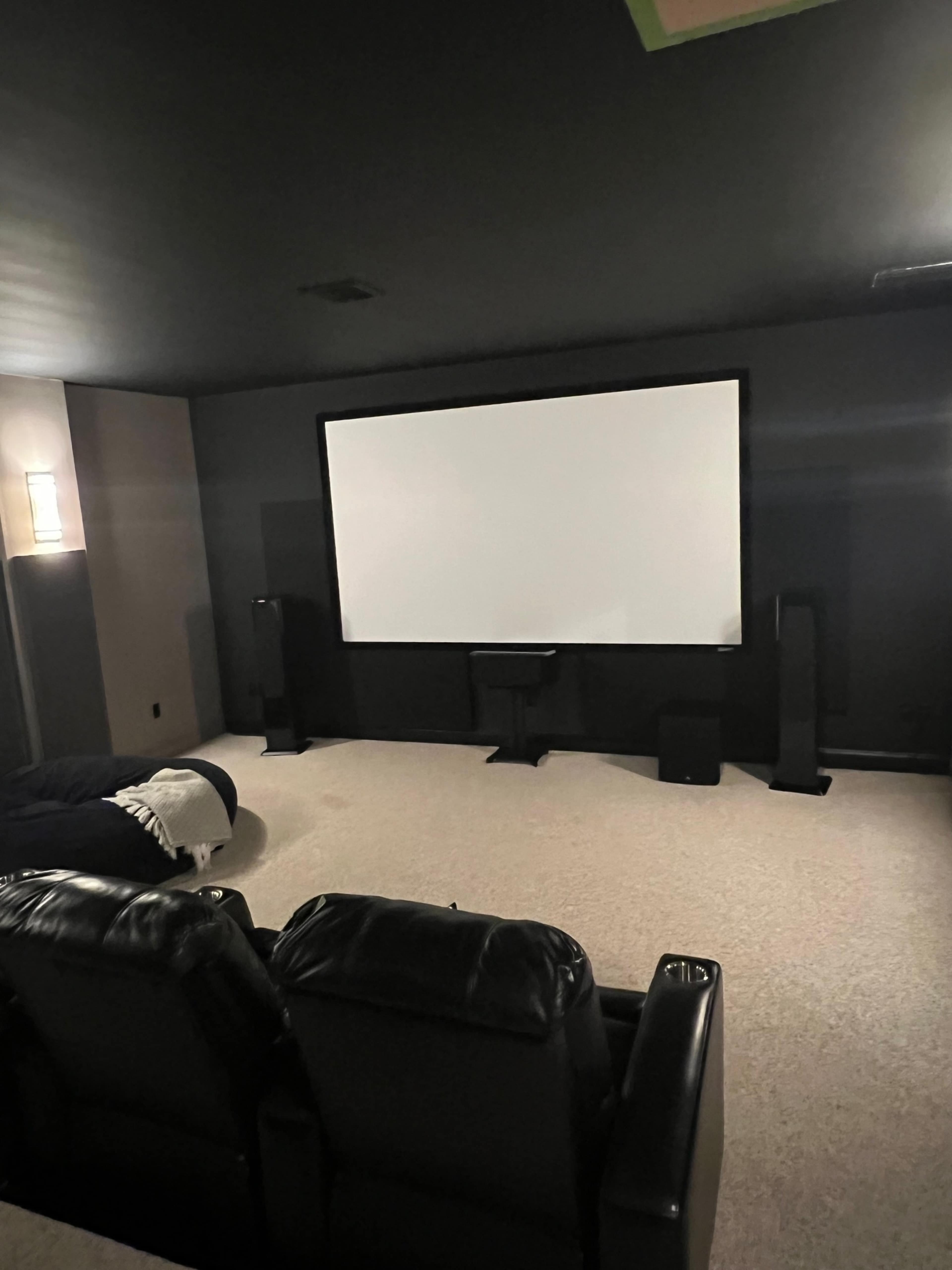 The image shows a dark home theater room with black seating facing a large blank screen and speakers positioned on either side.