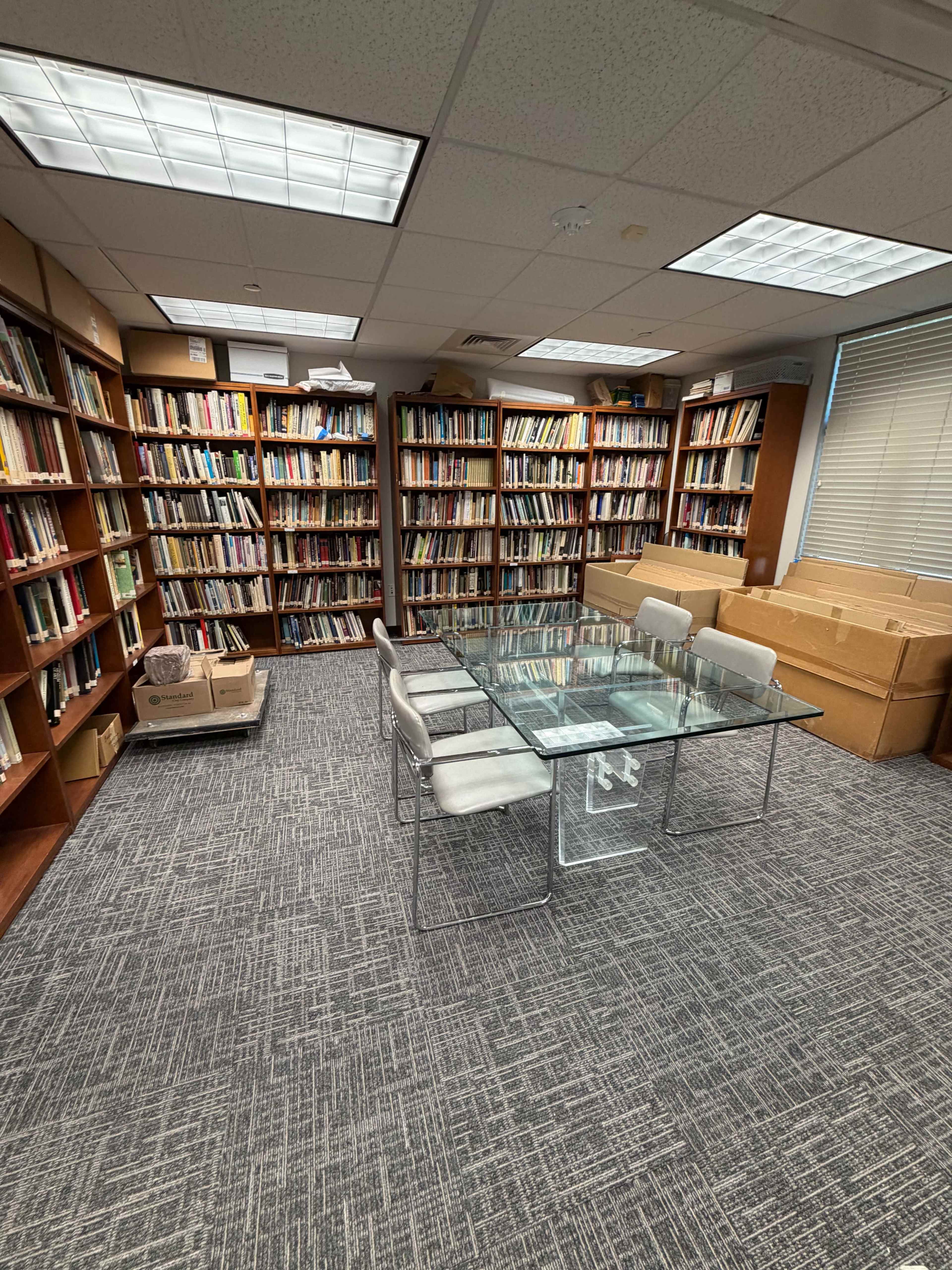 A glass table surrounded by four chairs sits in the center of a room filled with shelves of books, while cardboard boxes are stacked in one corner.