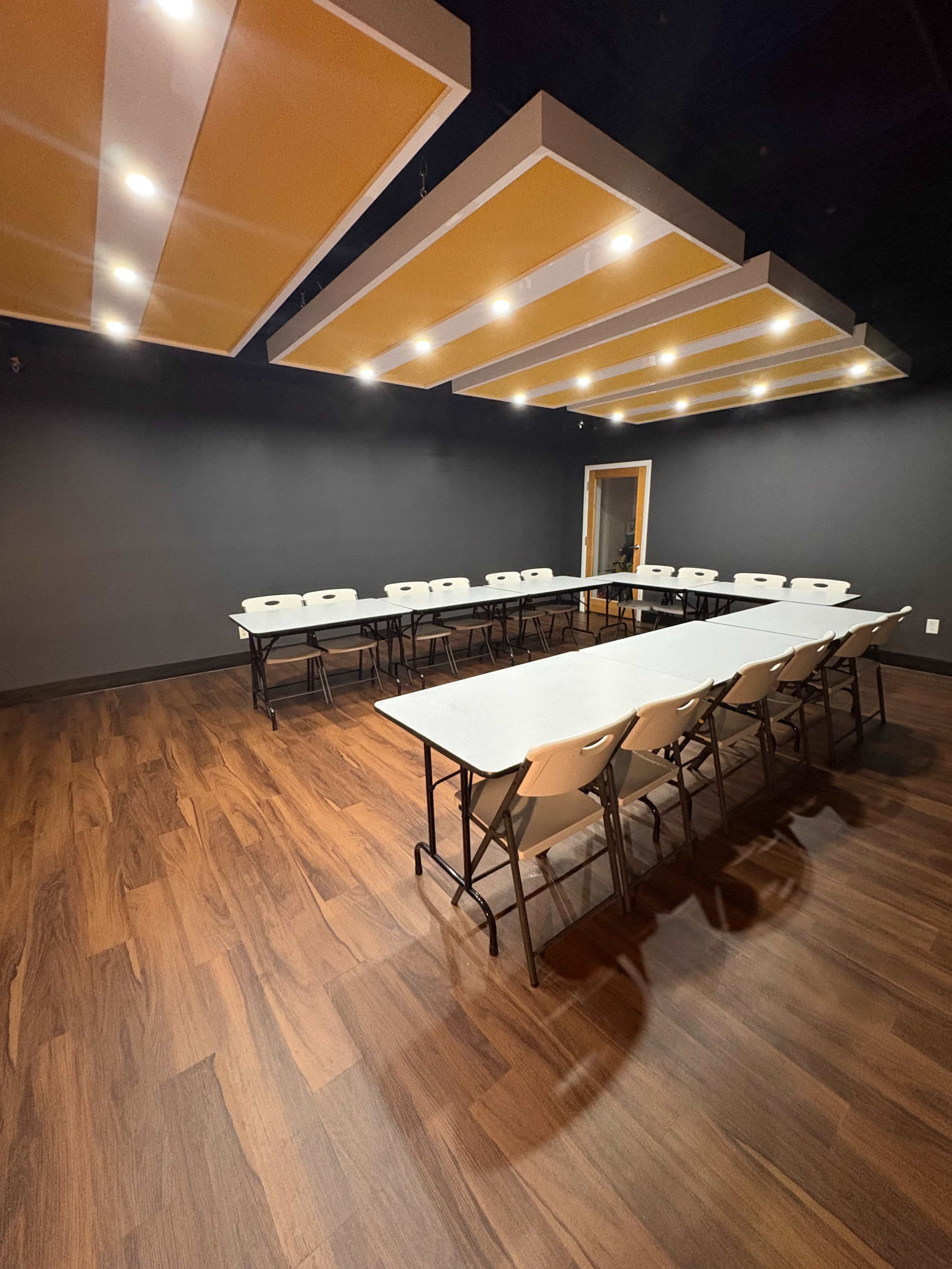 The image shows a meeting room arranged with folding tables and chairs in a U-shape, featuring a dark wall and lit ceiling panels.