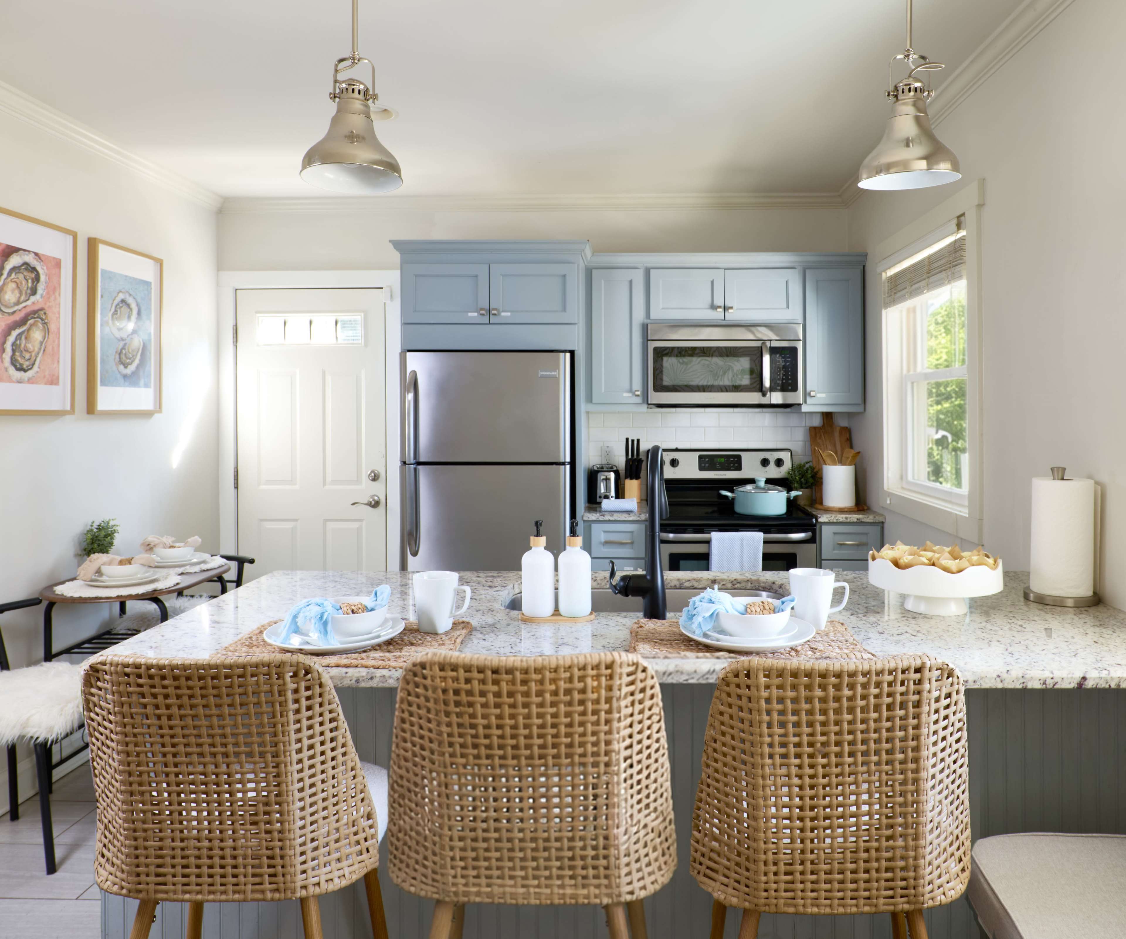 The image shows a modern kitchen with blue cabinets, stainless steel appliances, and a marble island featuring four wicker bar stools.