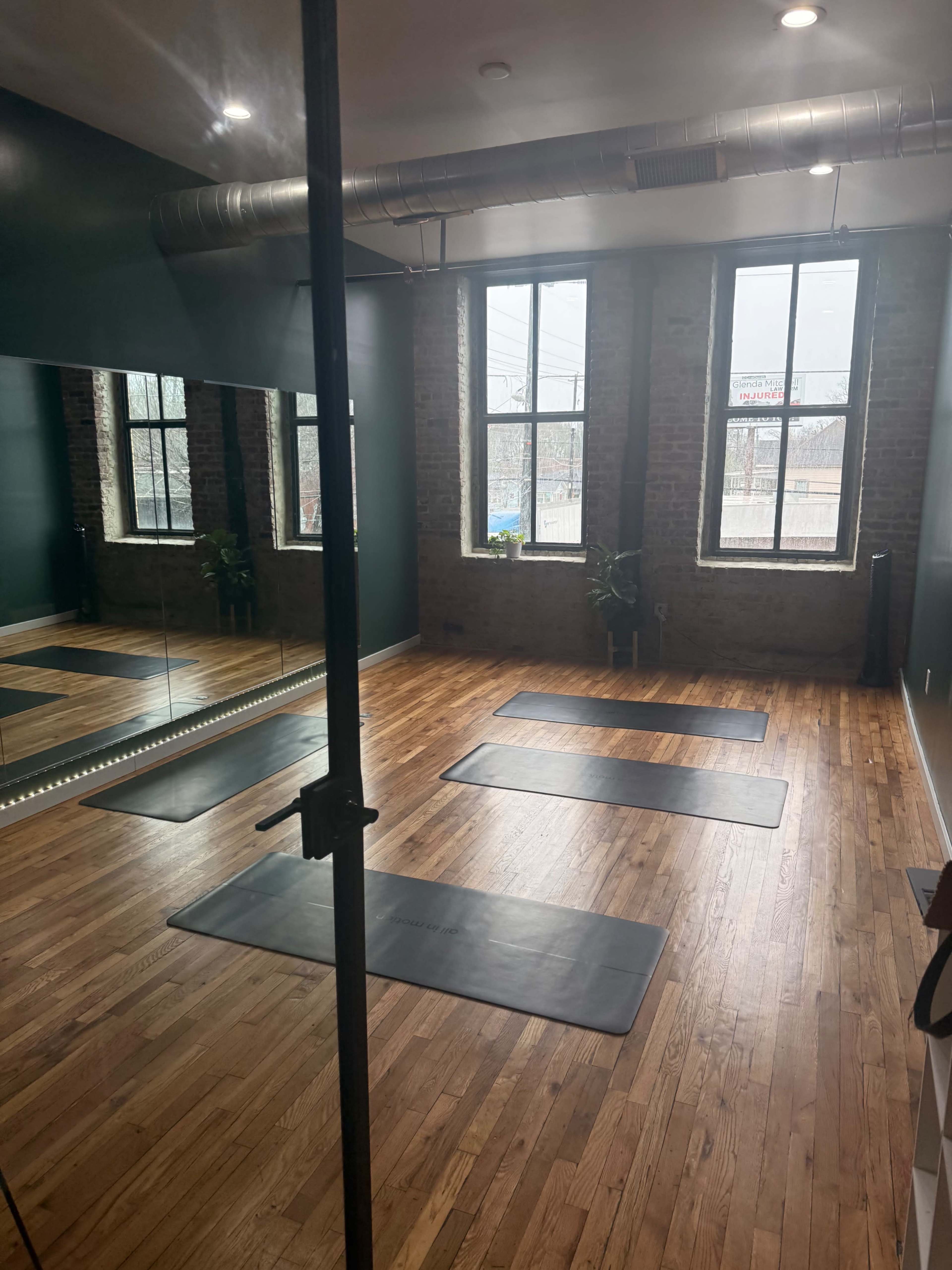 The image shows a spacious room with wooden floors, three yoga mats arranged on the floor, and large windows allowing natural light to filter in.