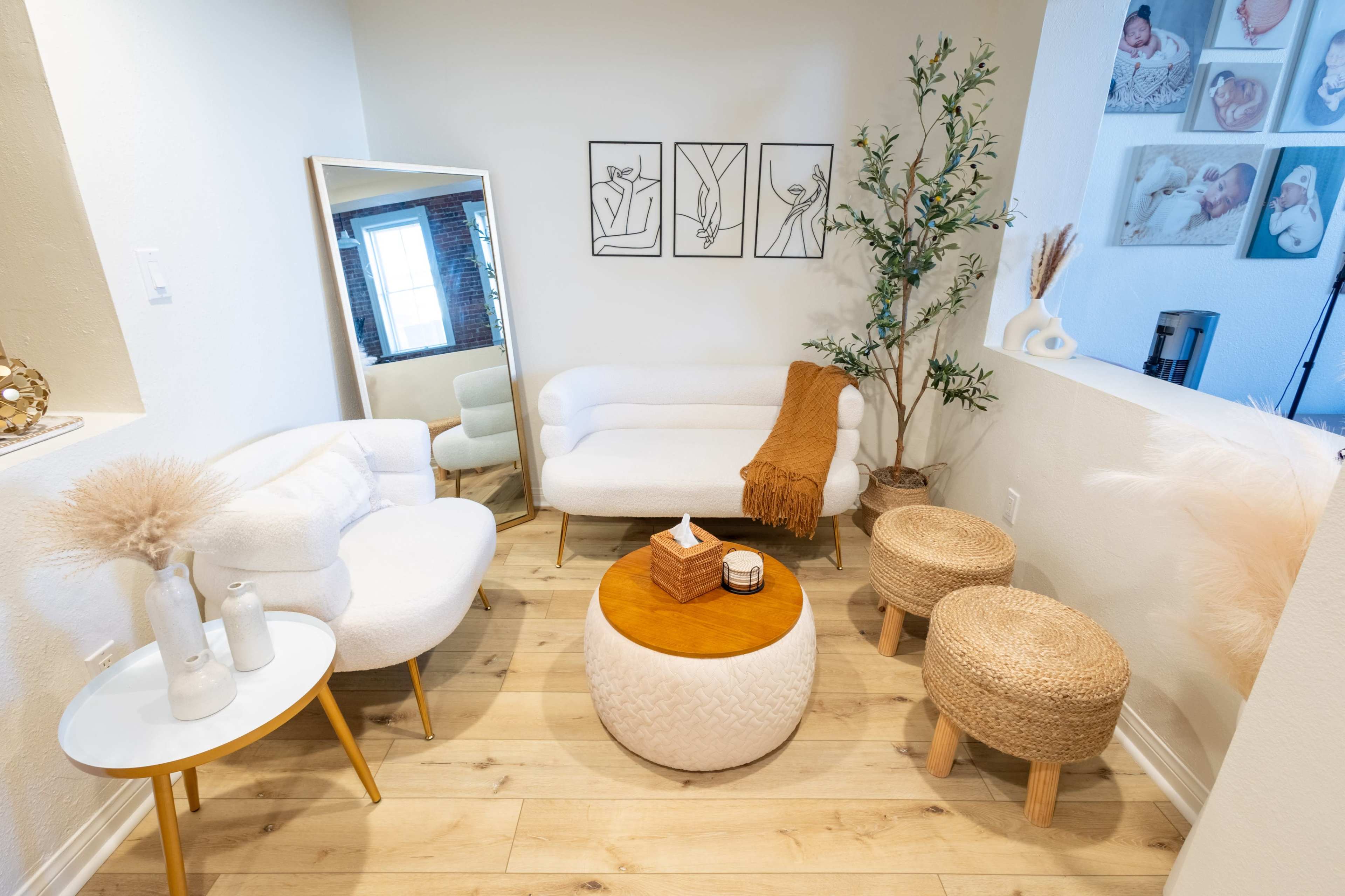 The image shows a cozy living room corner featuring two white sofas, a round wooden coffee table, and textured poufs, along with decorative plants and framed art on the walls.