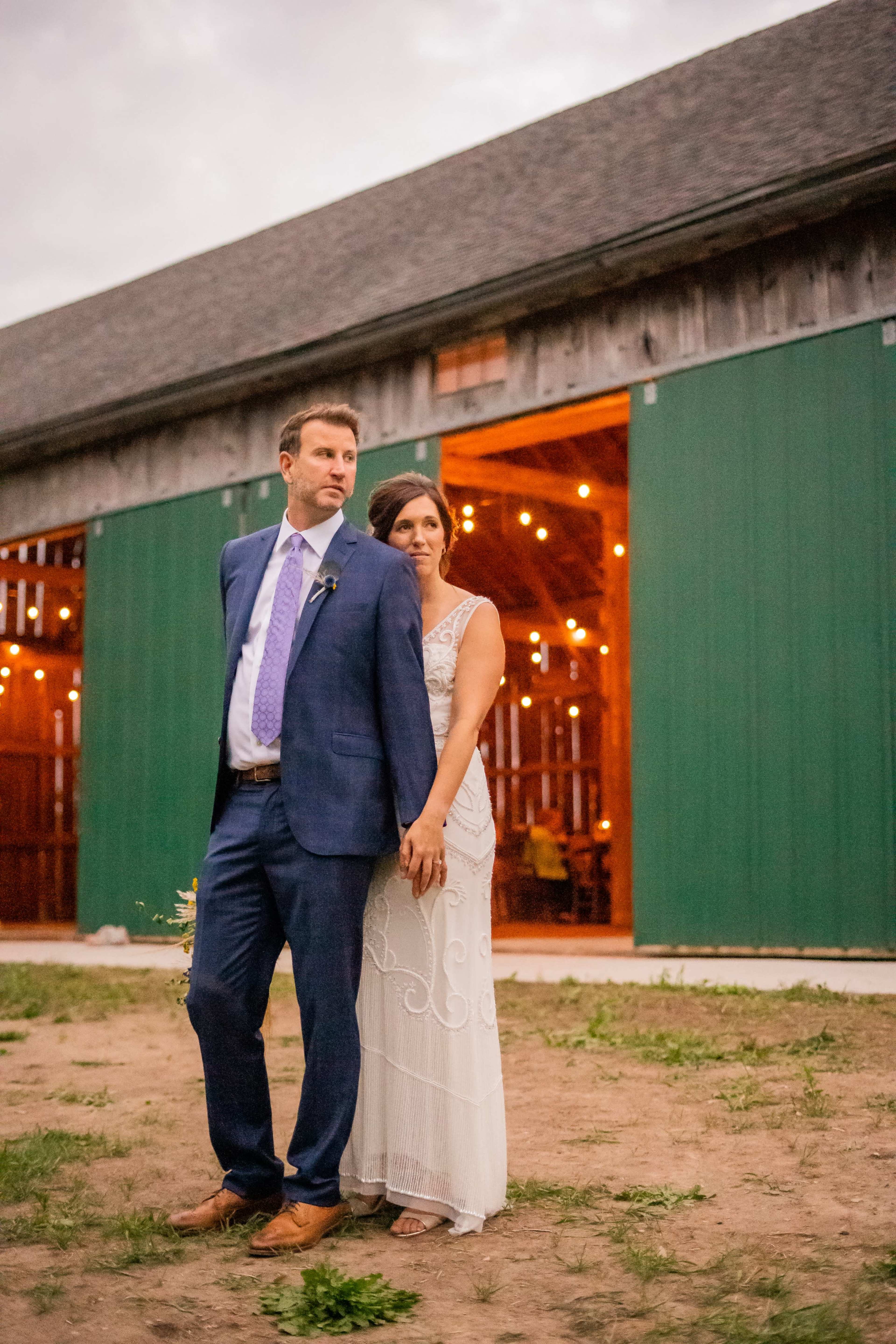 A couple stands together outside a barn, illuminated by soft string lights in the background.
