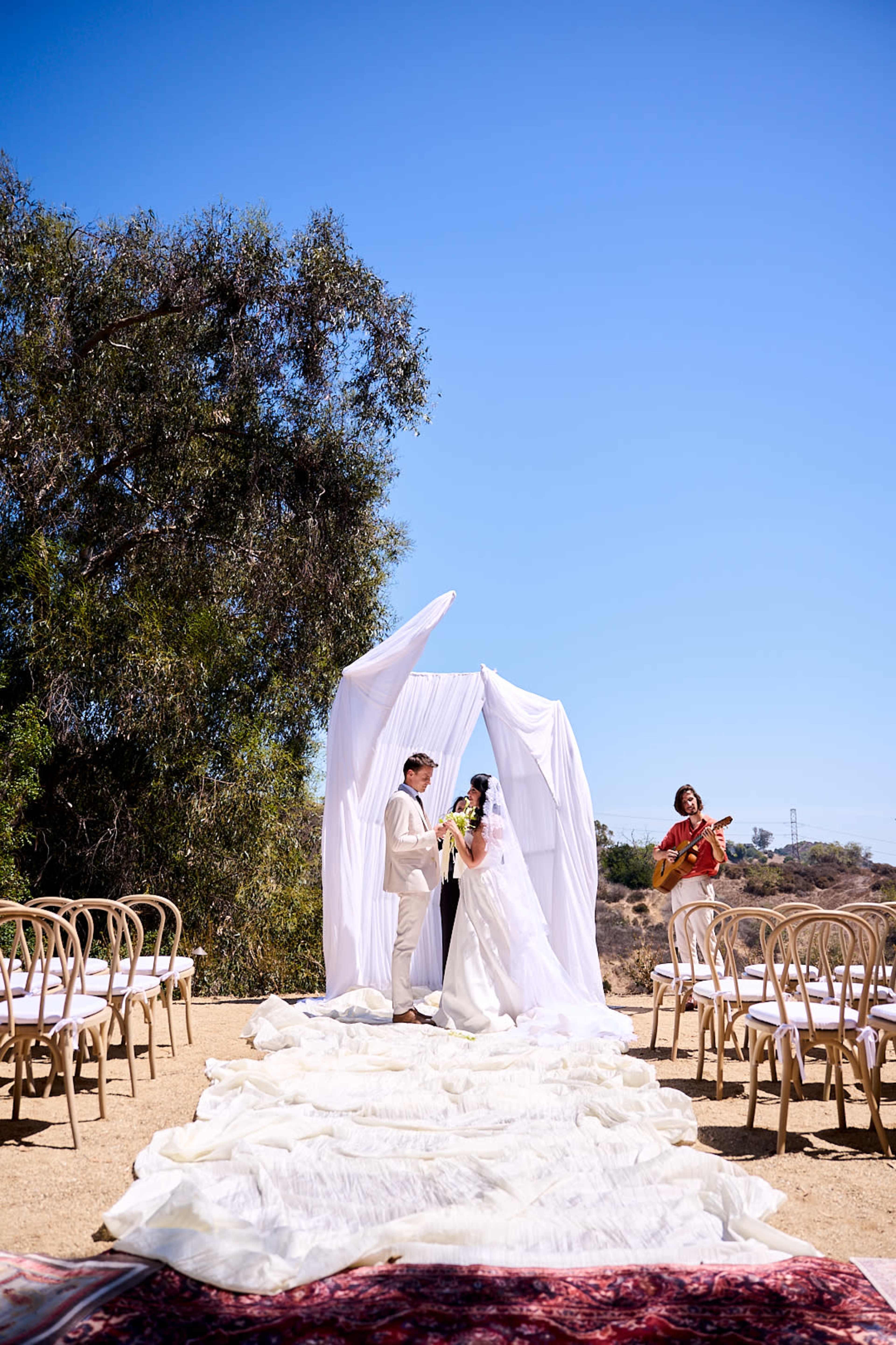 A couple stands beneath a white fabric arch during their wedding ceremony, while a musician plays guitar nearby and chairs are arranged on either side.