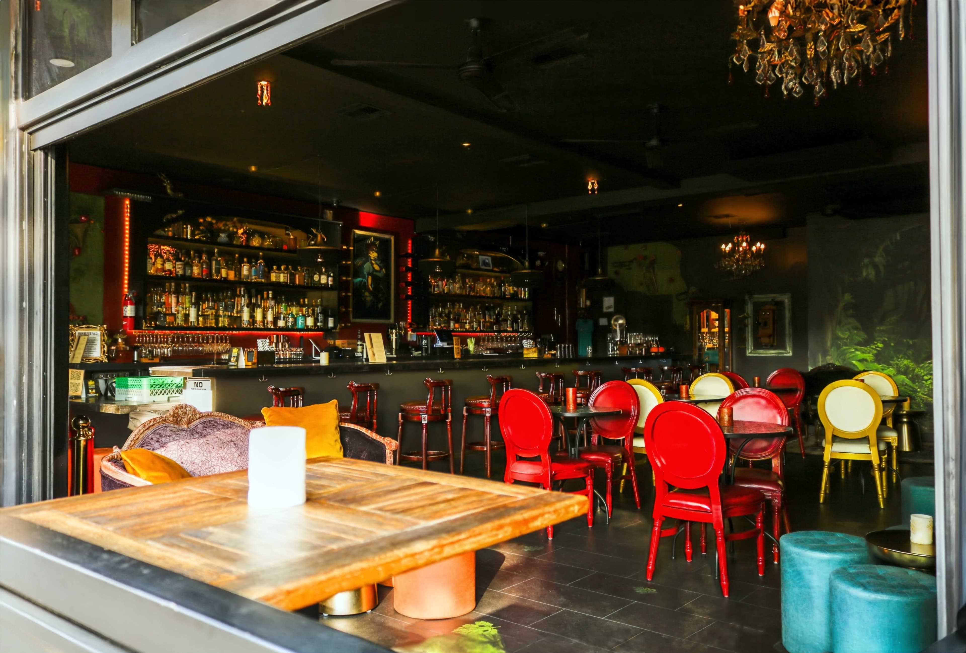 A bar area with a dark interior features a long shelf of bottles, red and gold chairs, and a wooden table in the foreground.