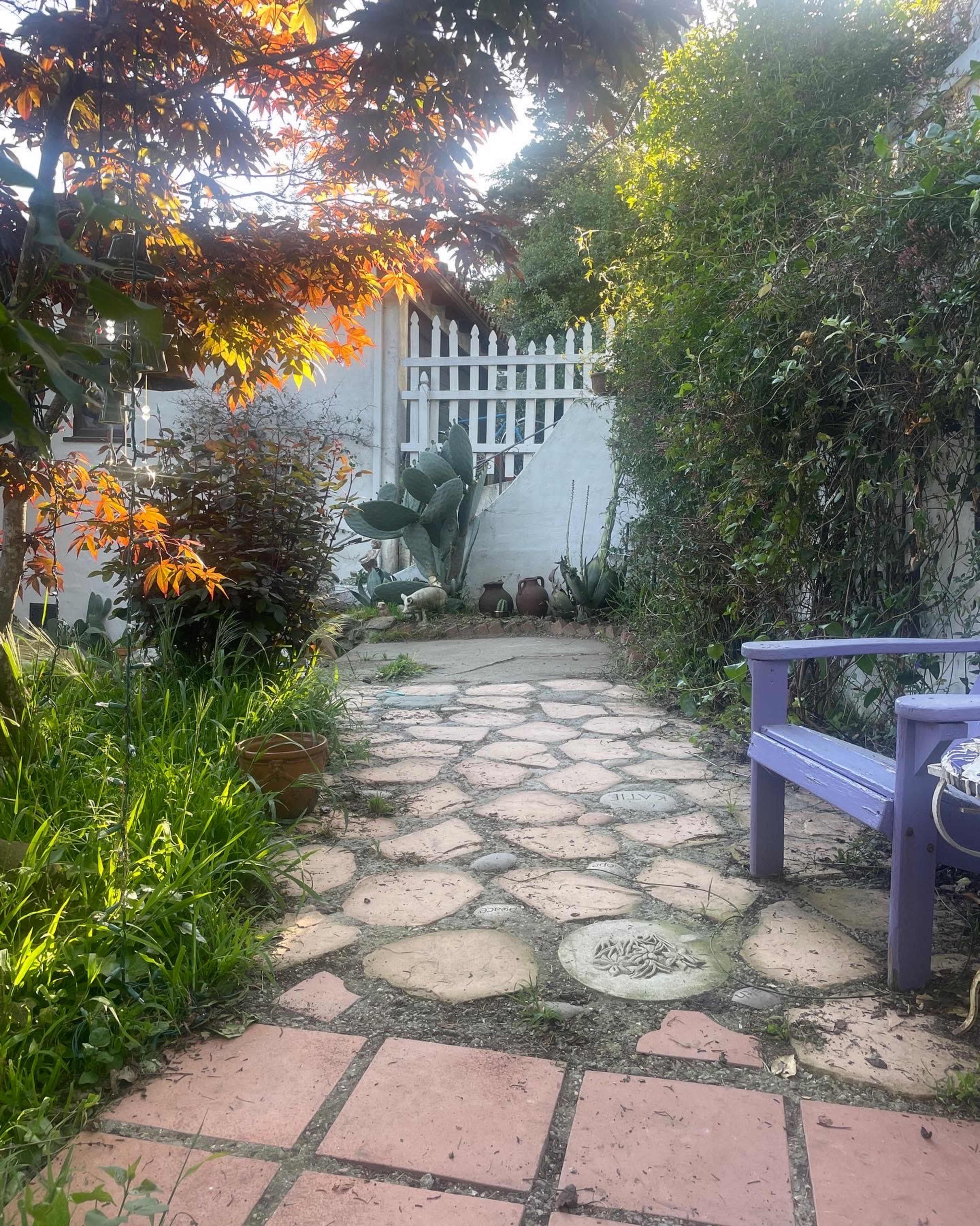 The image shows a stone pathway leading through a garden, bordered by lush greenery and a white wooden fence in the background.