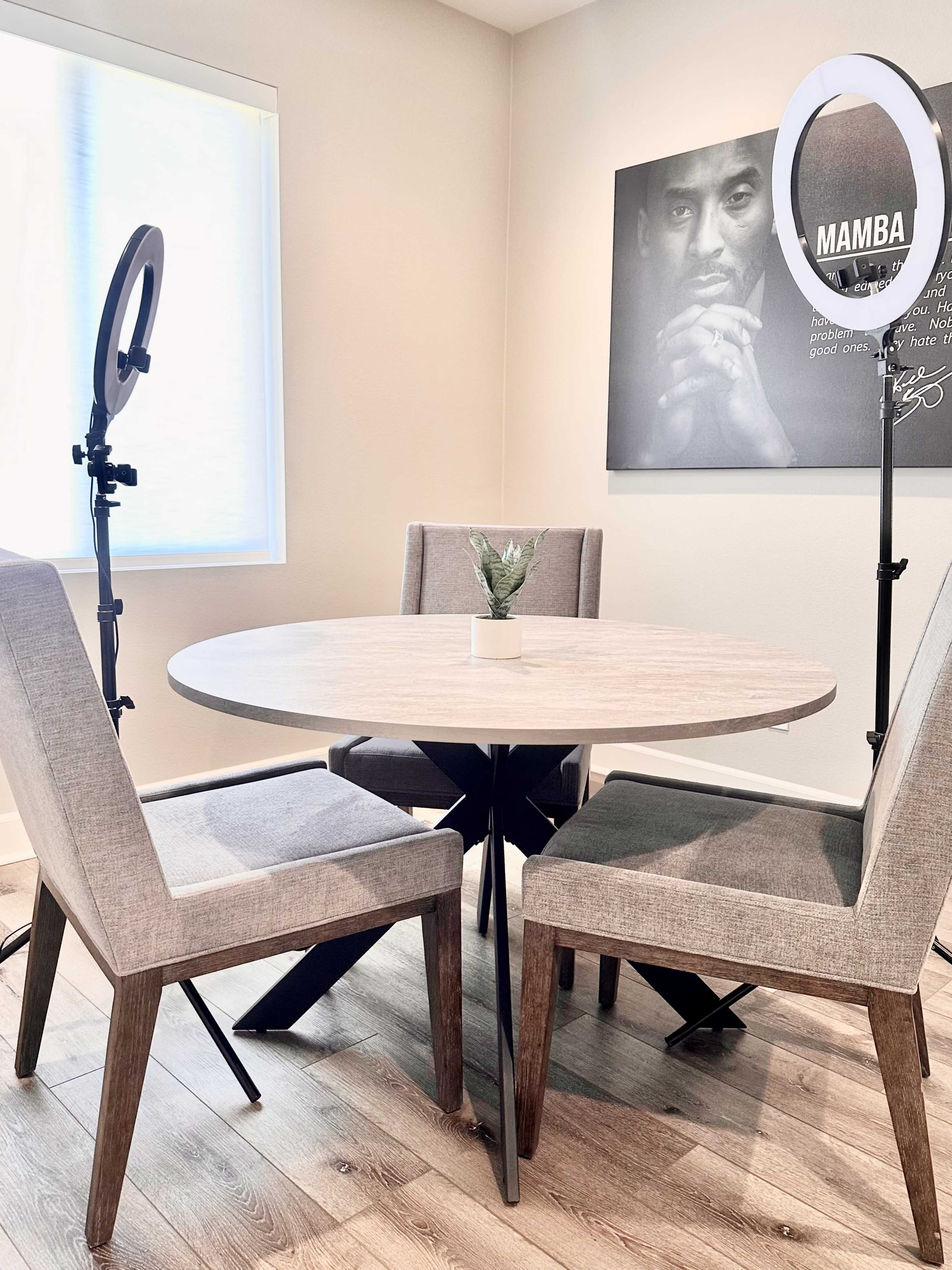 A round wooden table surrounded by three chairs, with two ring lights positioned on either side and a portrait of a man on the wall.