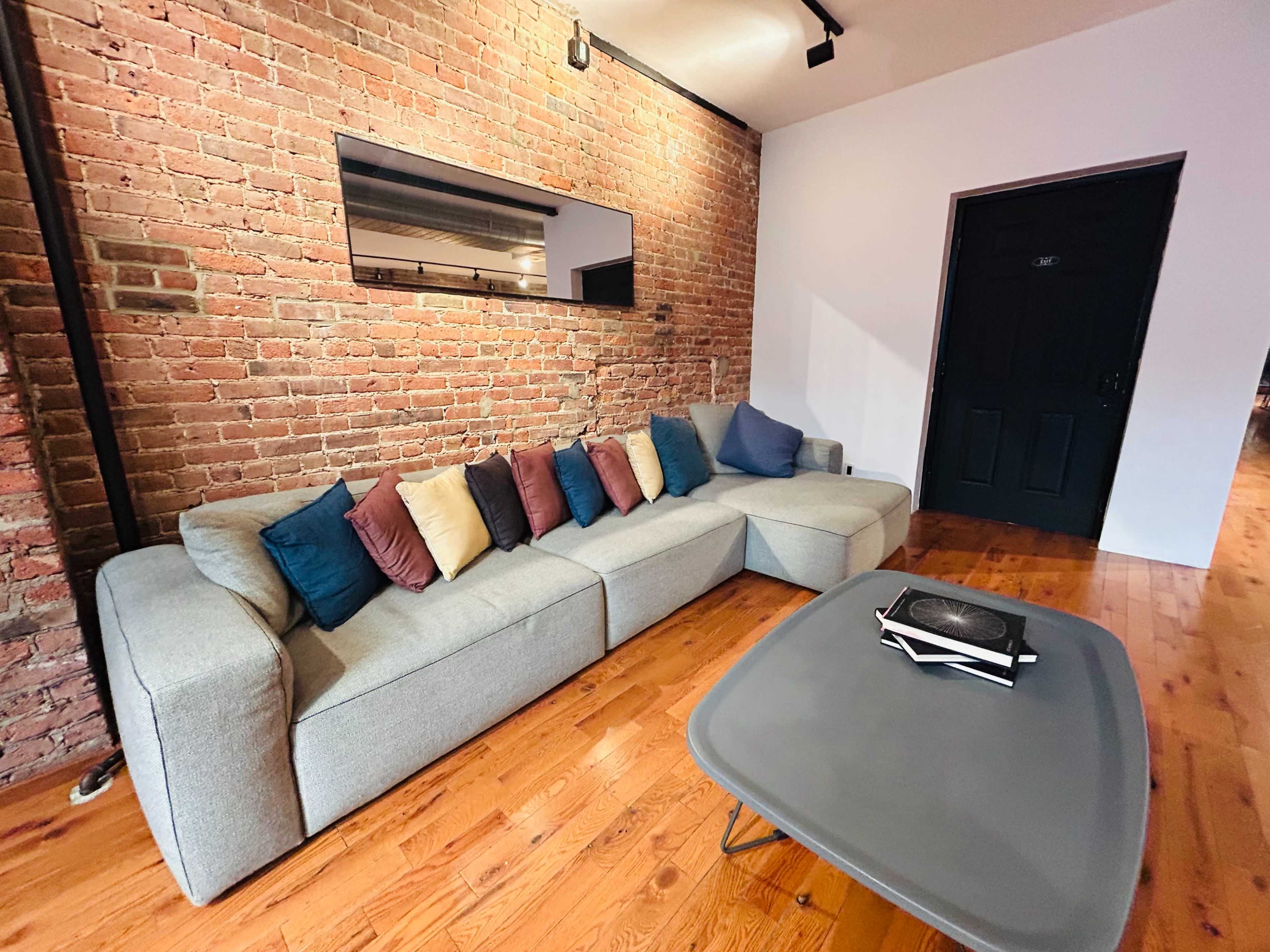 The image shows a modern living room with a gray sectional sofa adorned with colorful cushions, a wooden floor, and a brick wall with a mirror and dark door.