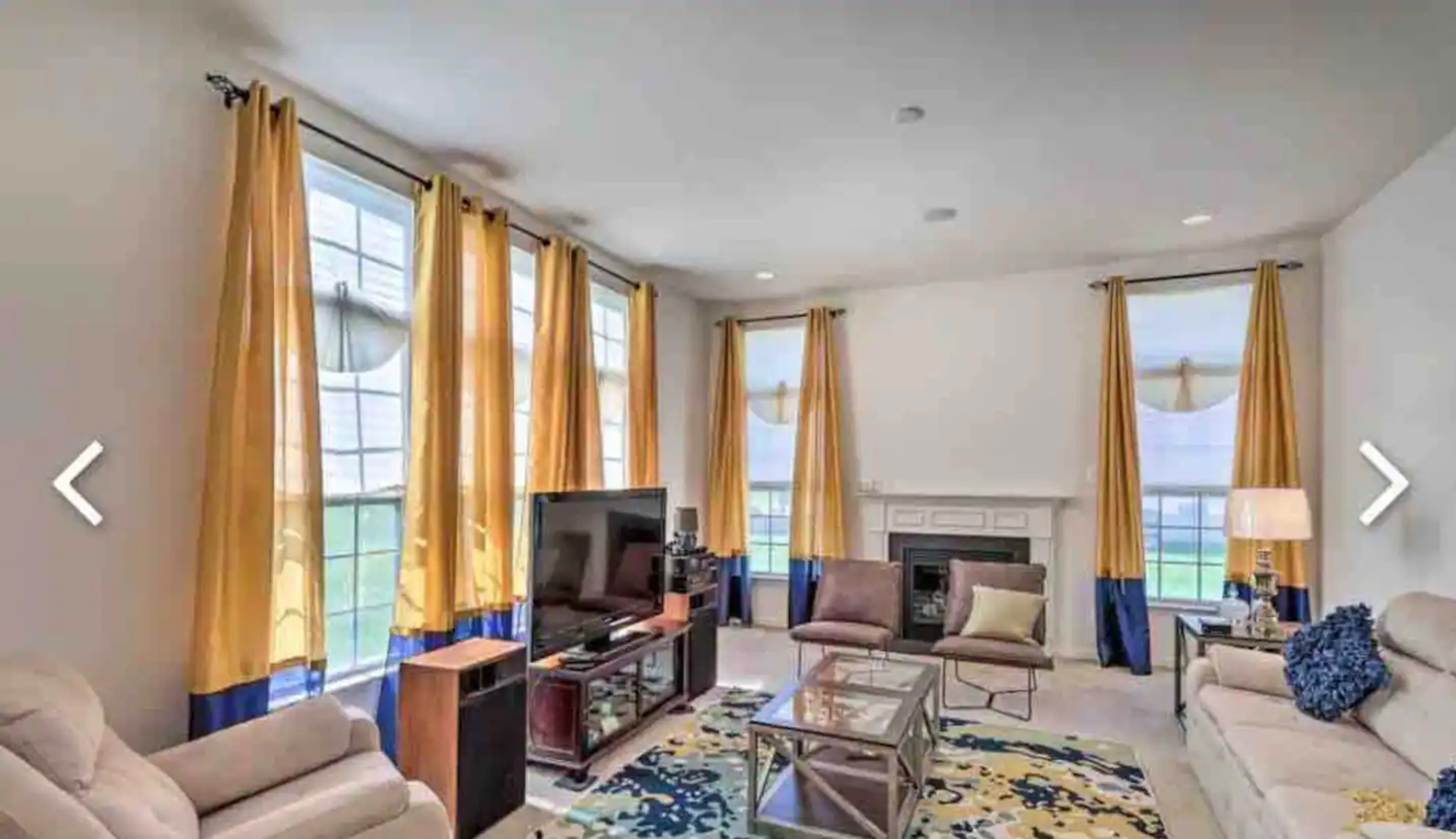 The living room features large windows dressed in yellow curtains, a television on a stand, a glass coffee table, and beige sofas arranged around a central area.