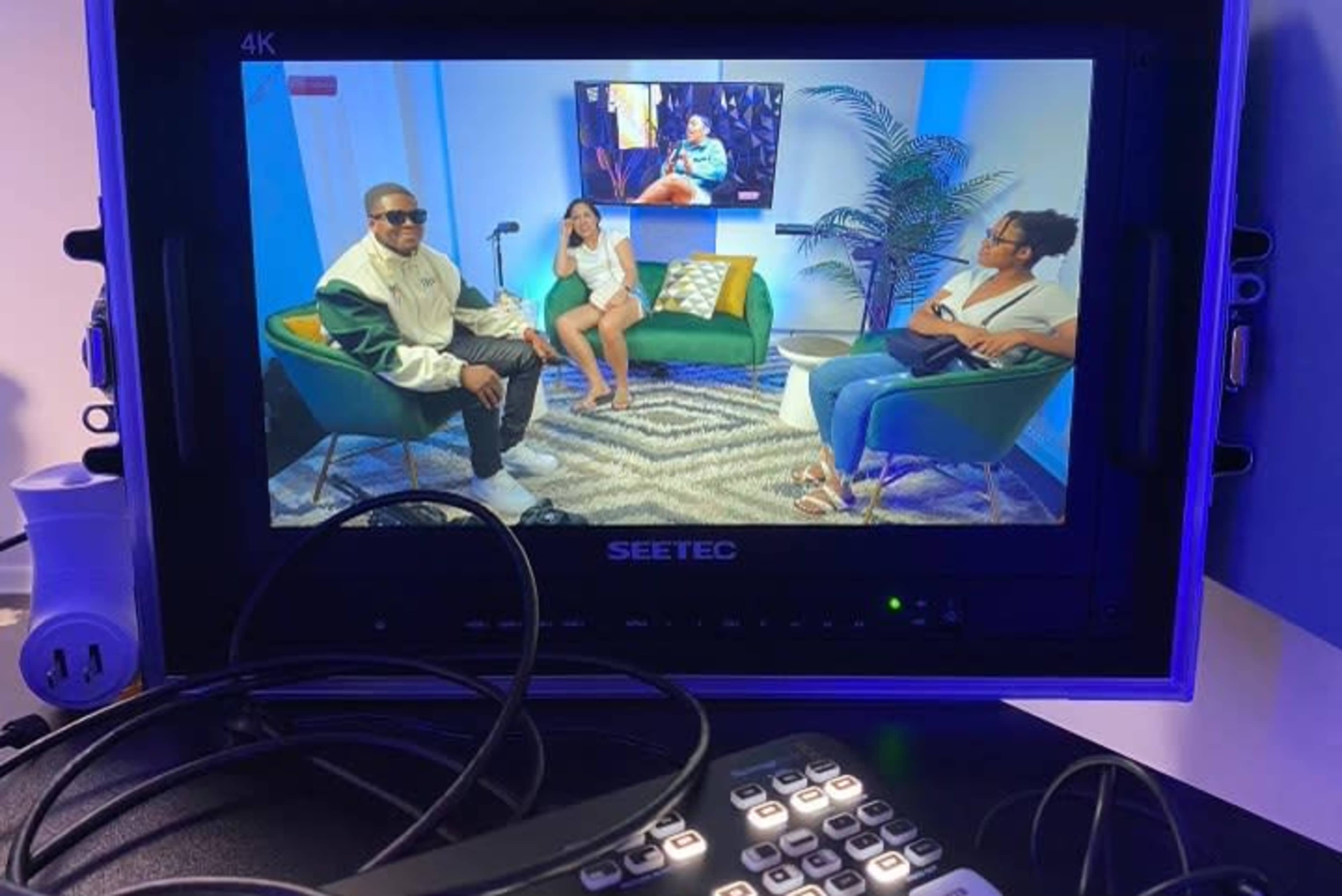 A television monitor displaying a talk show set with three people seated in green chairs, surrounded by a plant and a colorful rug.