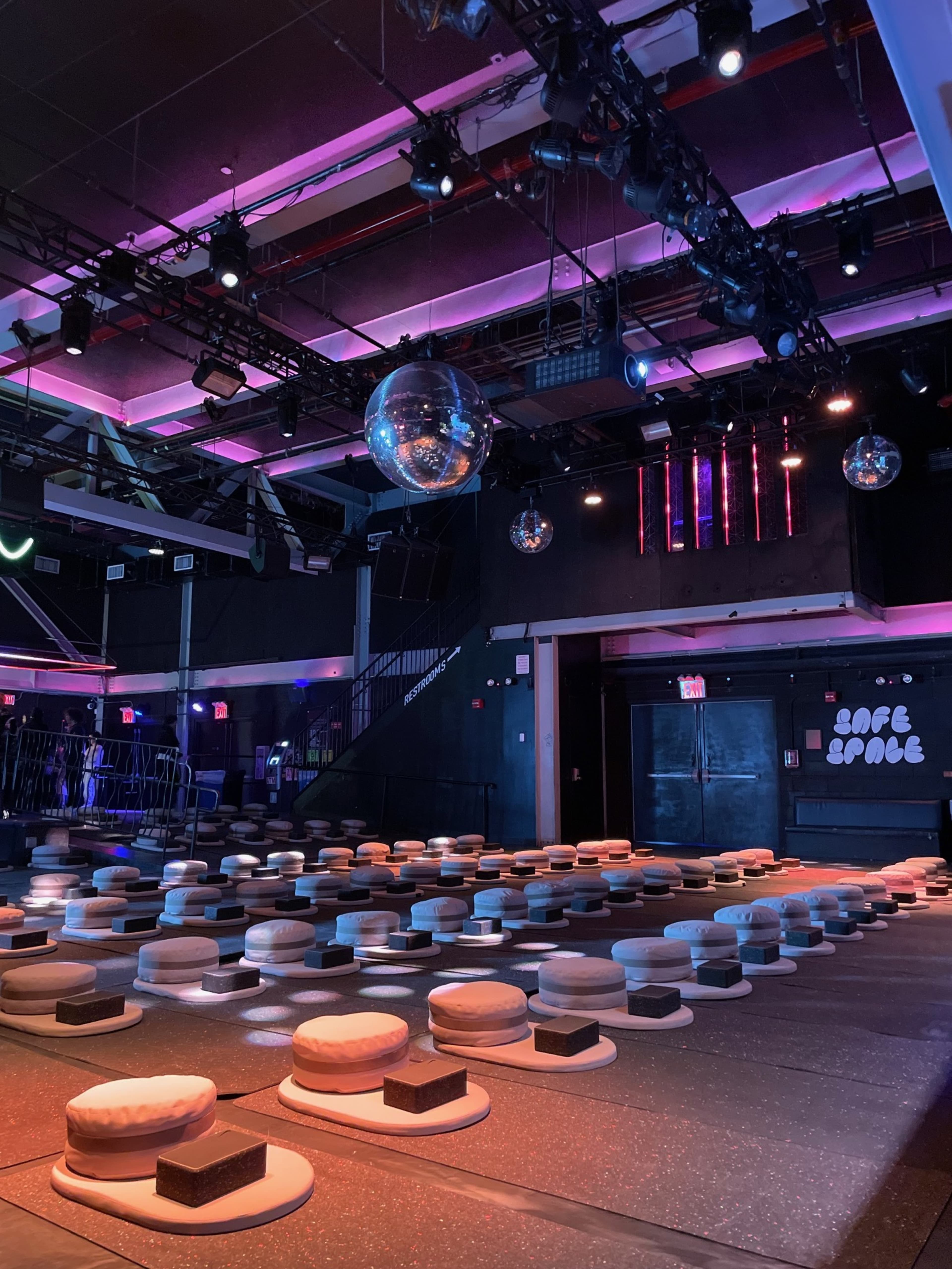 The image shows a dimly lit performance space set up with rows of round cushions on a dark floor, illuminated by colorful stage lights and featuring a disco ball overhead.