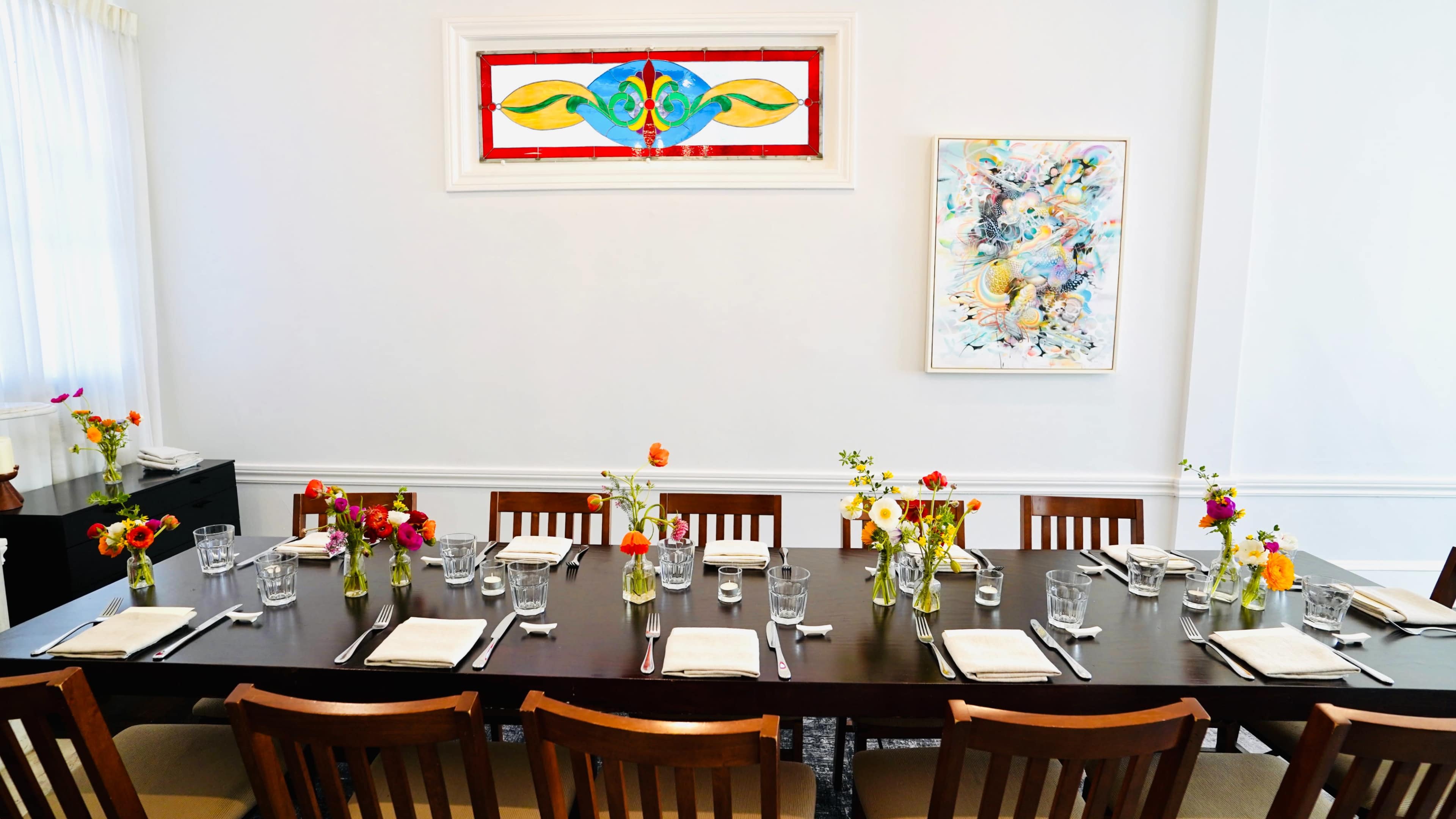 A long dining table is set with glasses and floral arrangements, surrounded by wooden chairs in a bright room with a stained glass window and a colorful artwork on the wall.