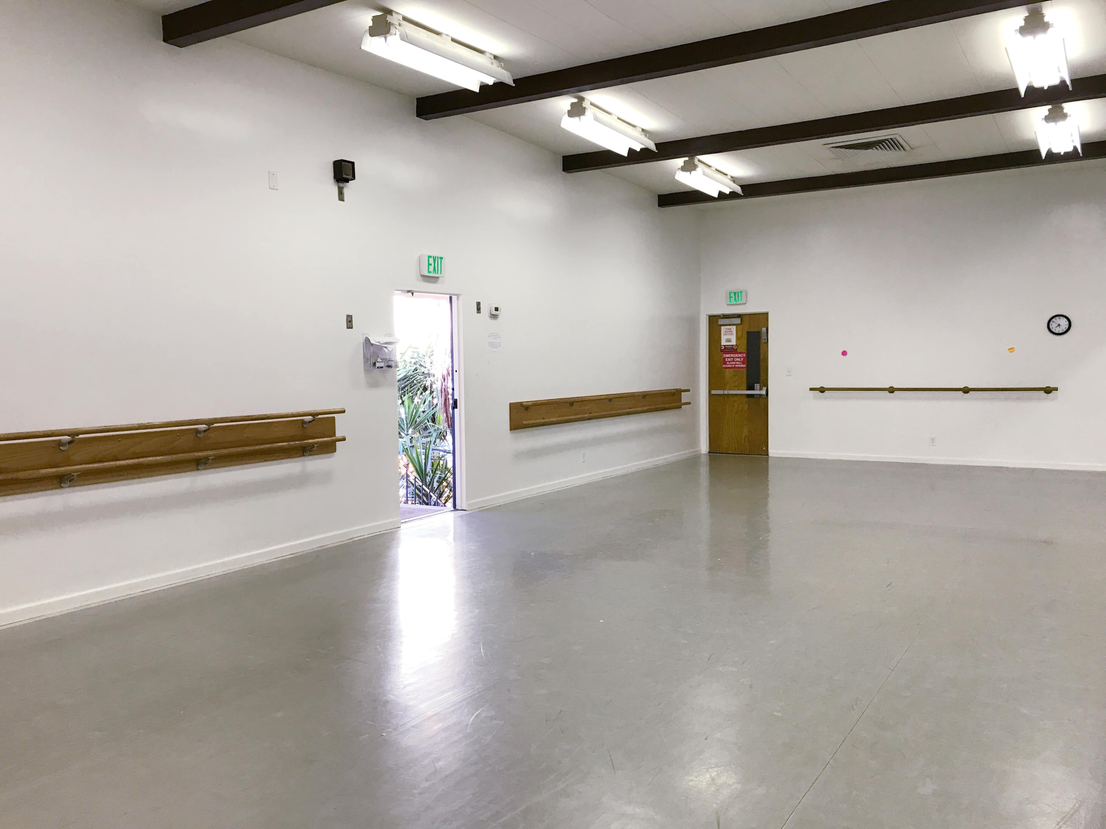 An empty dance studio features mirrored walls, wooden ballet bars, and a door opening to an outdoor area.
