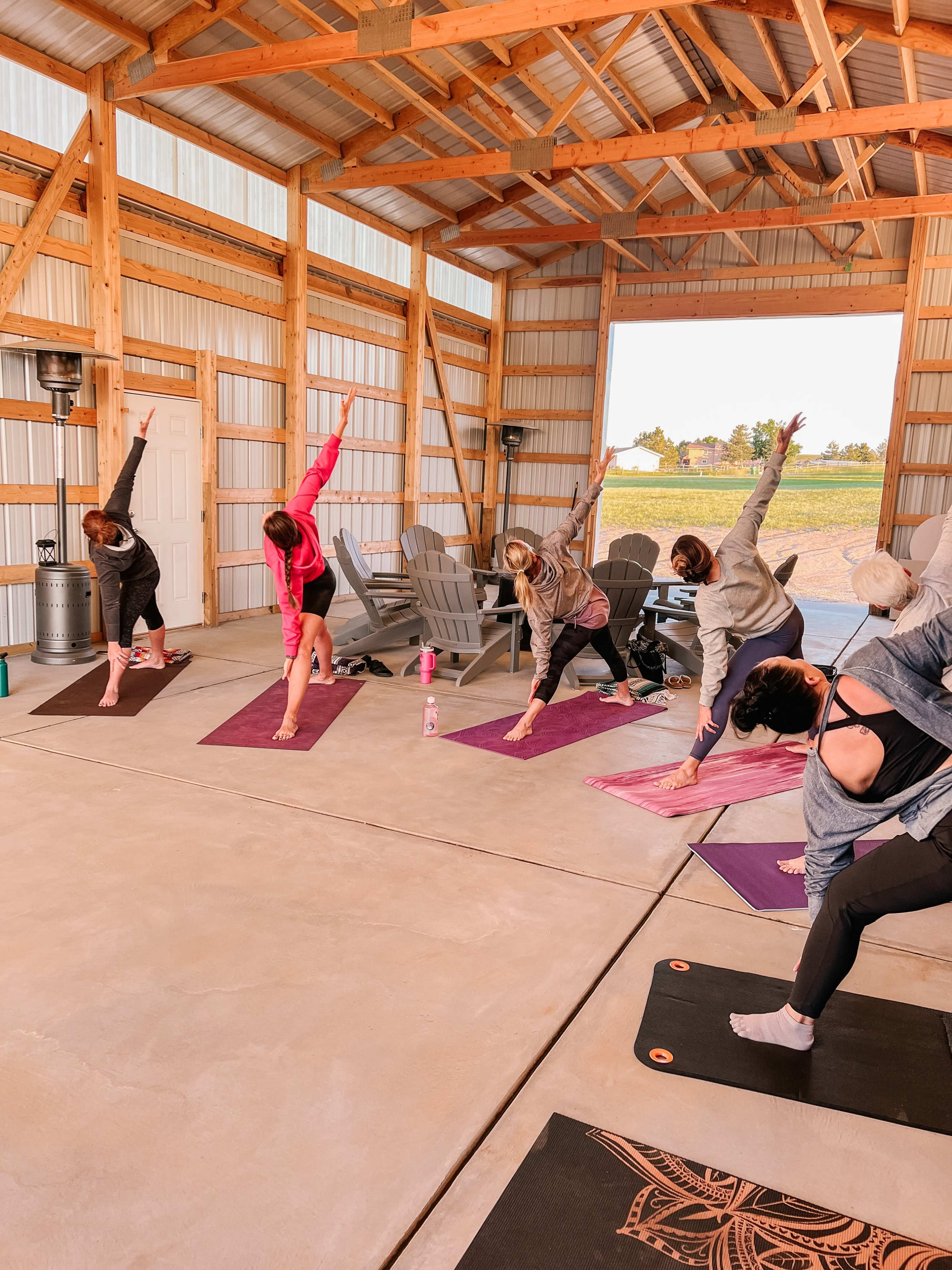 A group of individuals is practicing yoga in a spacious, open building with wooden beams, using yoga mats on a concrete floor.