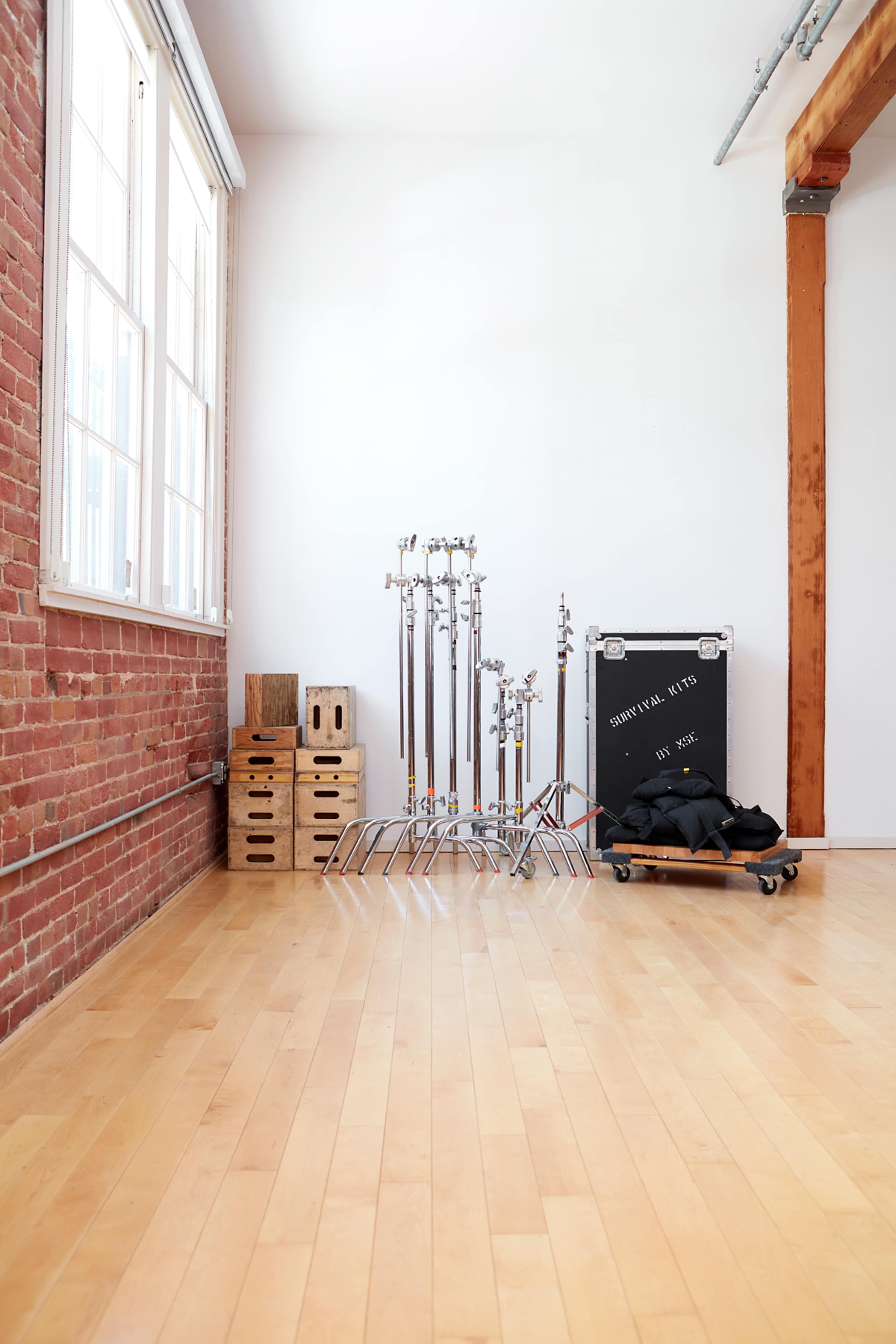 A spacious room with wooden flooring features a wall of red brick, storage crates, and various lighting equipment stands near a black storage case.