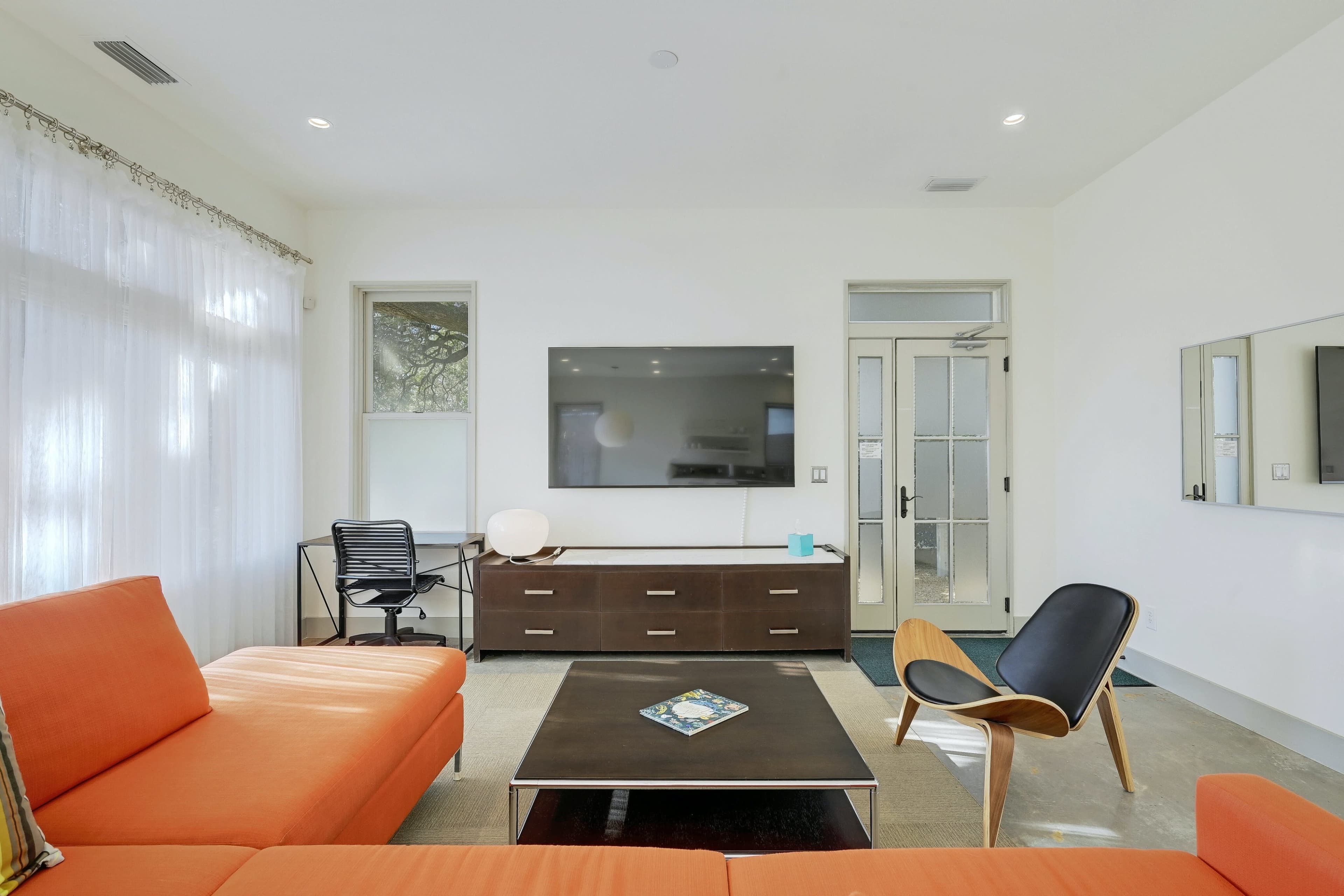 A modern living room features an orange sectional sofa, a black chair, a coffee table, and a desk beside a glass door.
