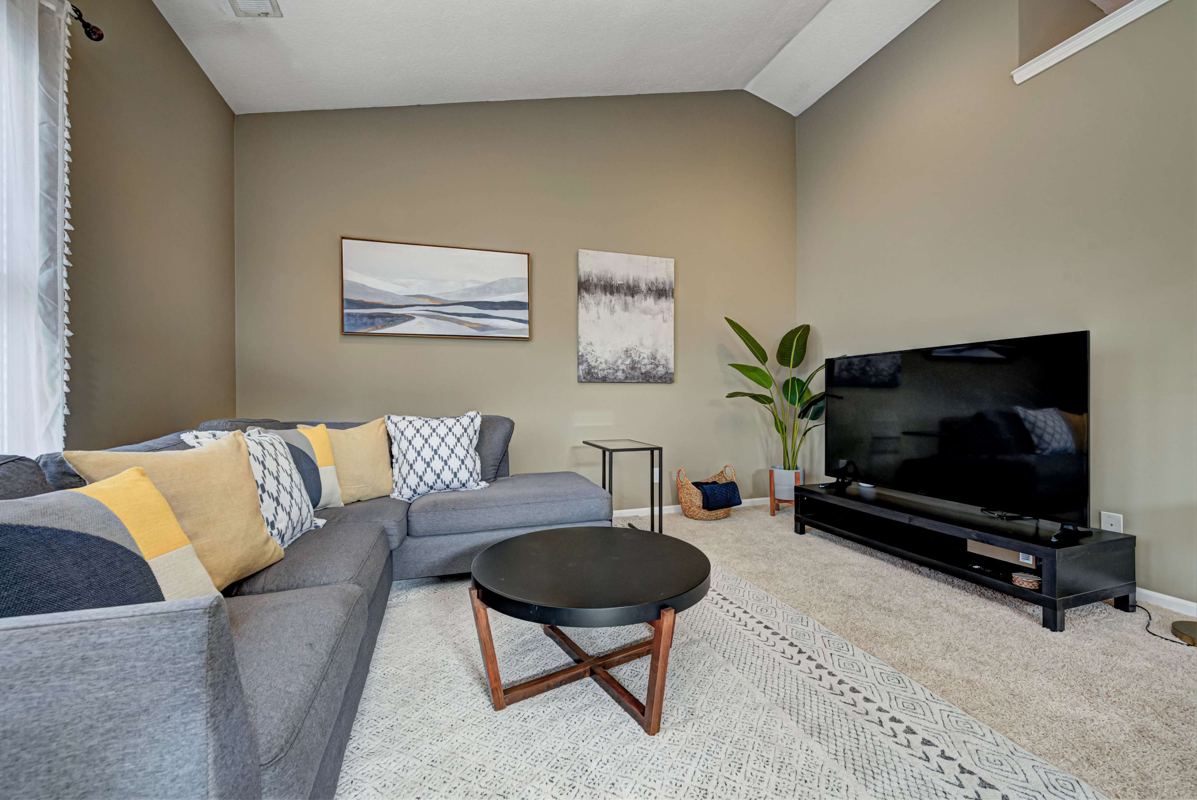 A living room with a sectional sofa, a round coffee table, a television stand, and two framed artworks on the walls.