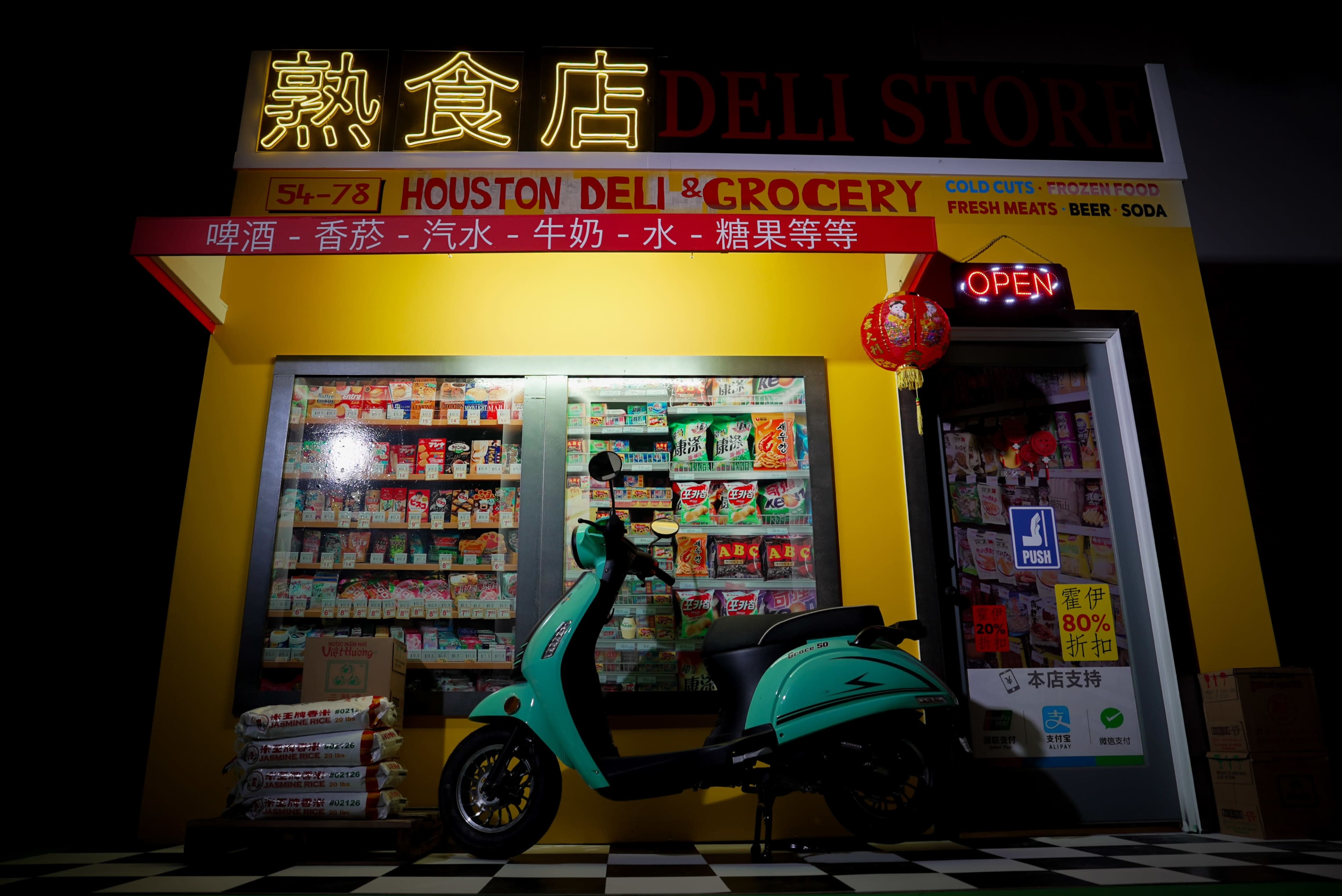 A brightly lit deli store featuring a turquoise scooter in front of shelves stocked with various groceries and a red lantern hanging above the entrance.