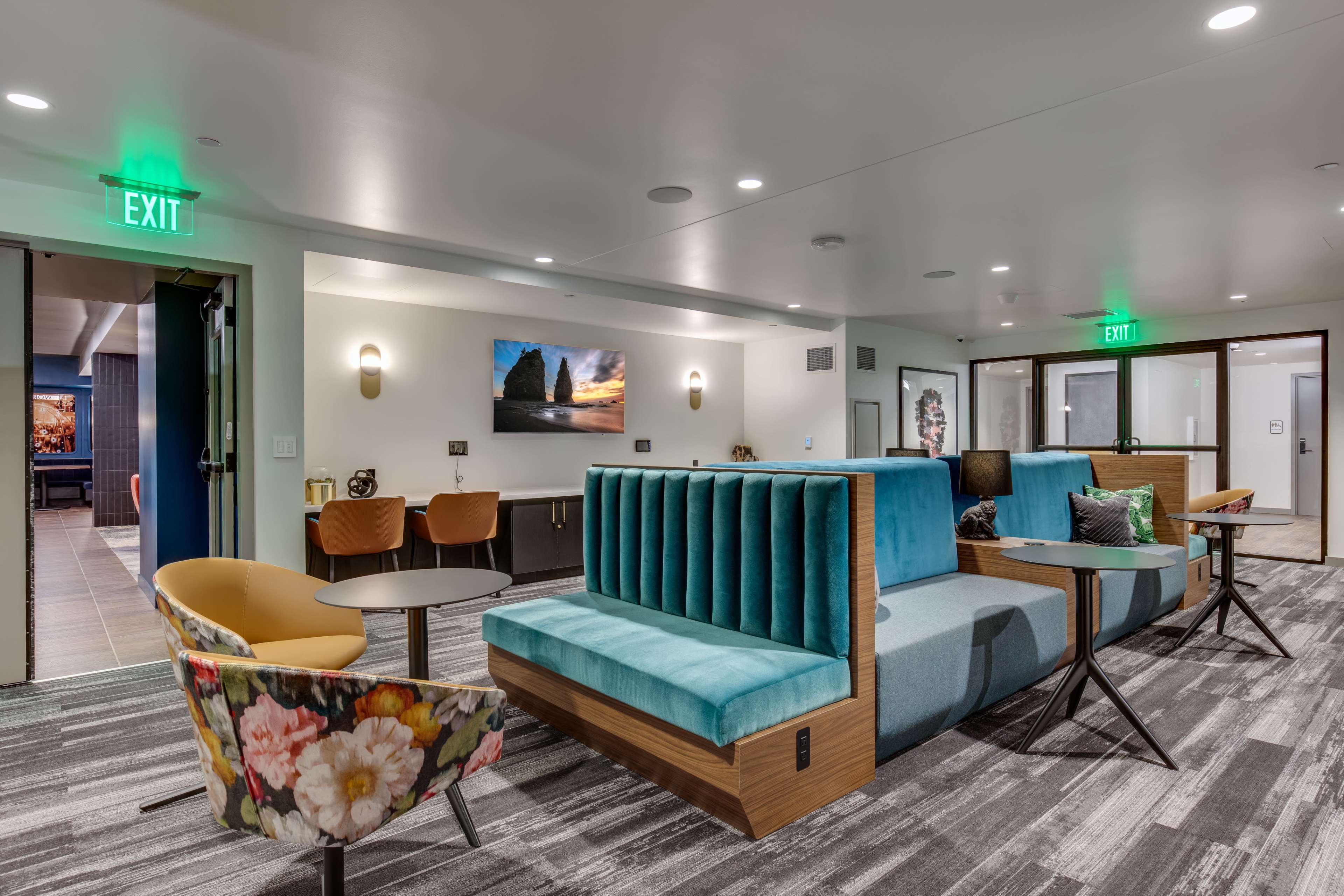 A modern lounge area featuring a large turquoise sectional sofa, a table, and colorful chairs against a backdrop of muted walls and patterned flooring.