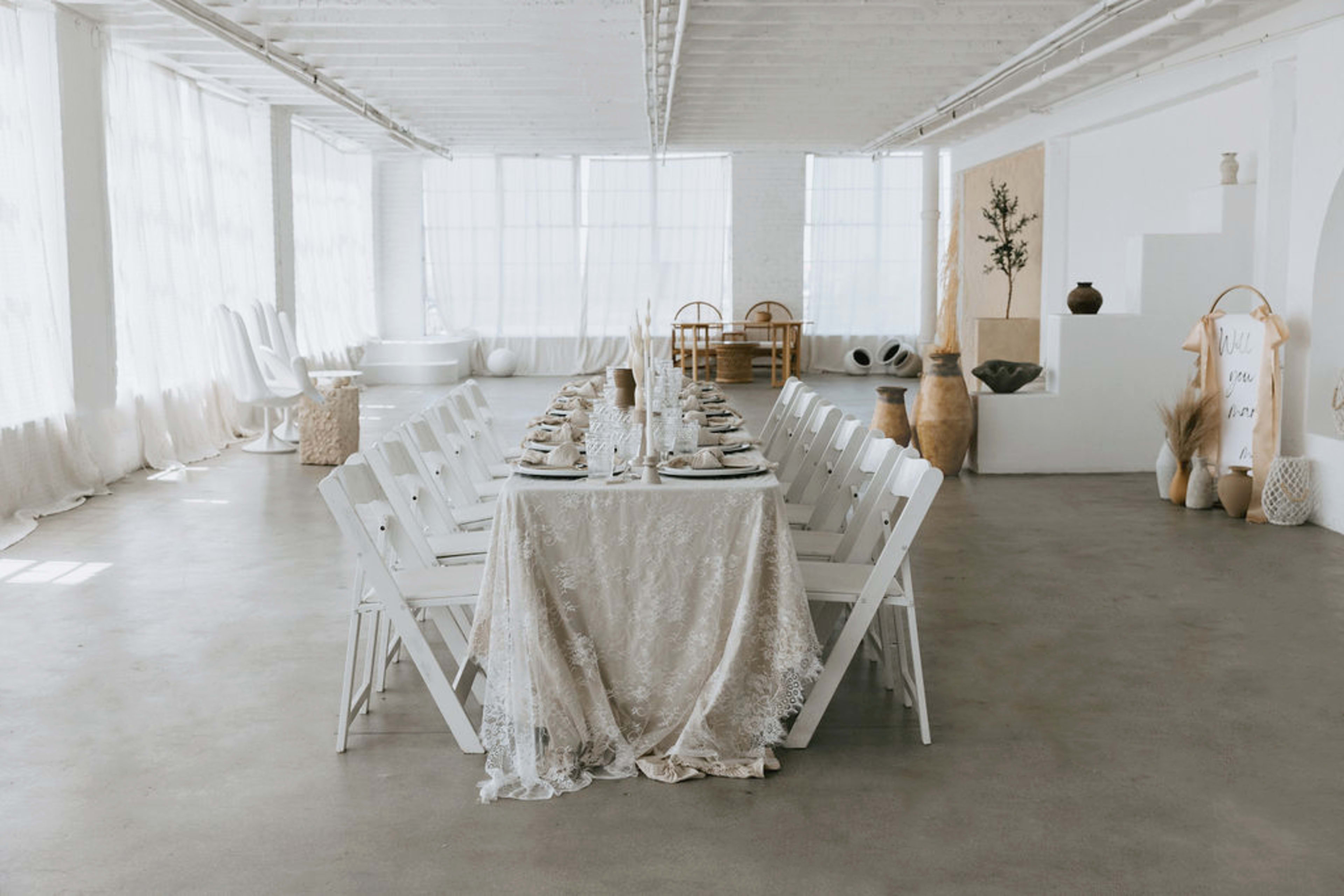 A long, elegantly set dining table with white chairs is arranged in a bright, airy room featuring large windows and minimalistic decor.