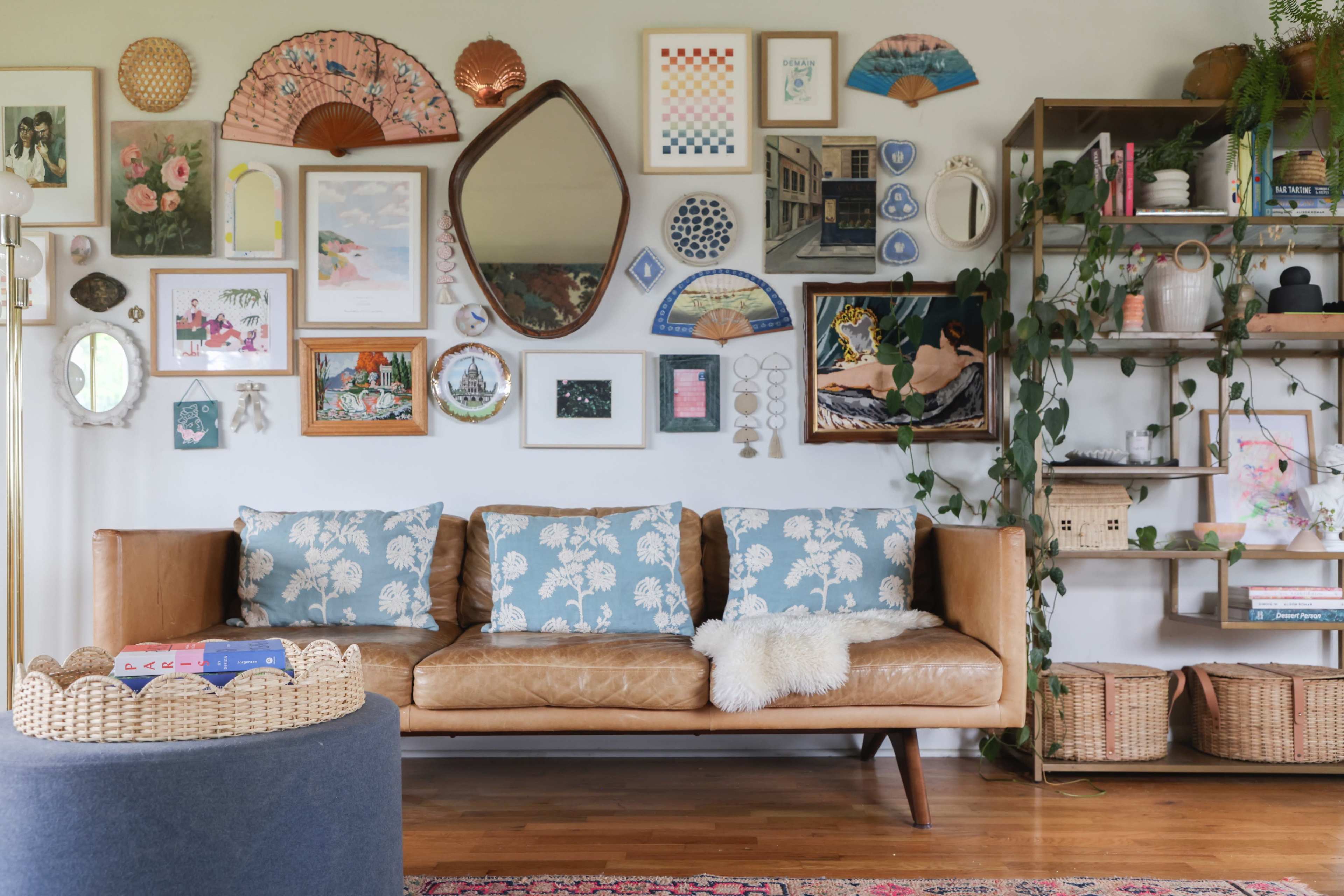 A mid-century modern living room features a brown leather sofa with patterned cushions, surrounded by an eclectic gallery wall and a shelf filled with decorative items and plants.