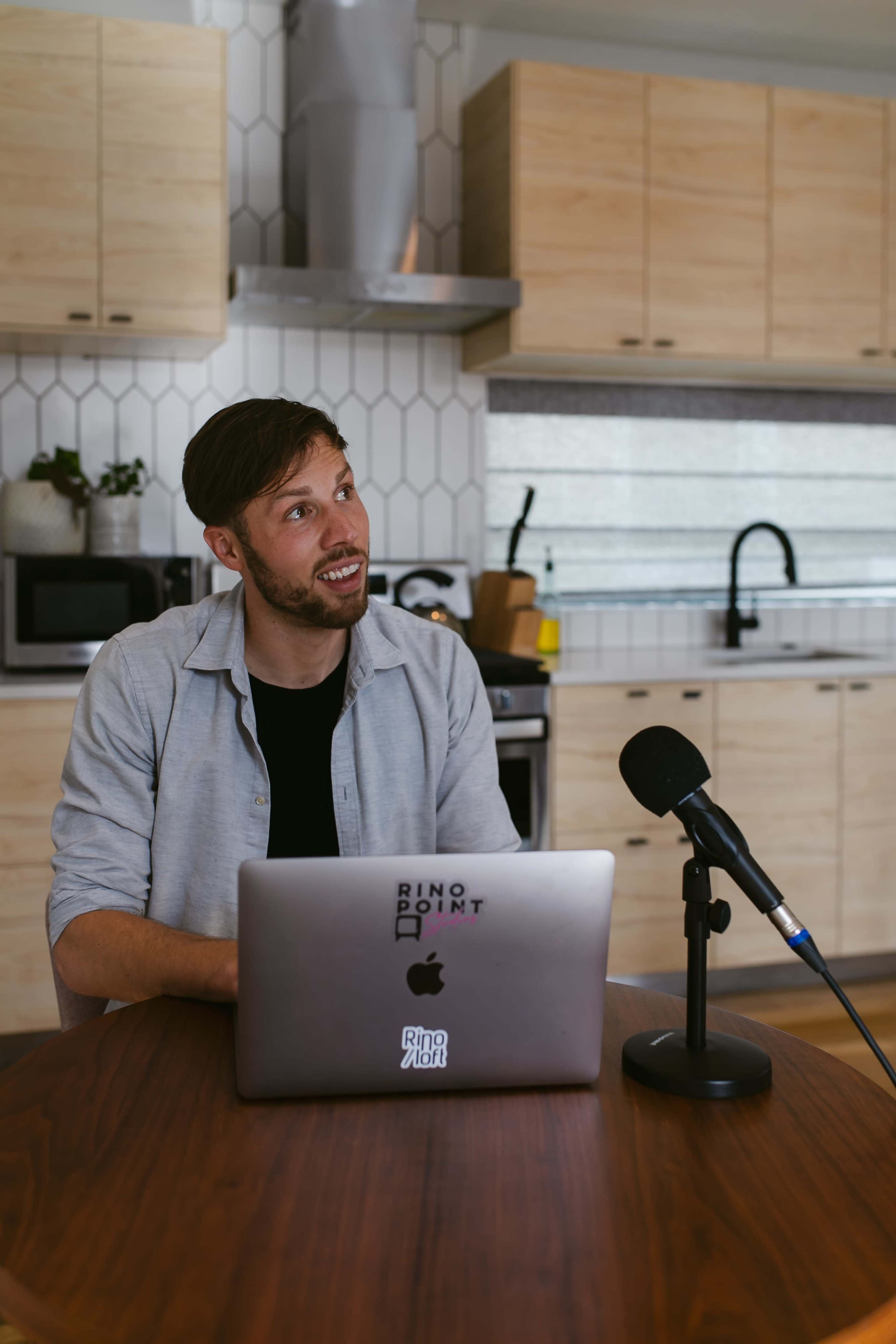 A man sits at a wooden table with a laptop open in front of him and a microphone beside him in a modern kitchen.