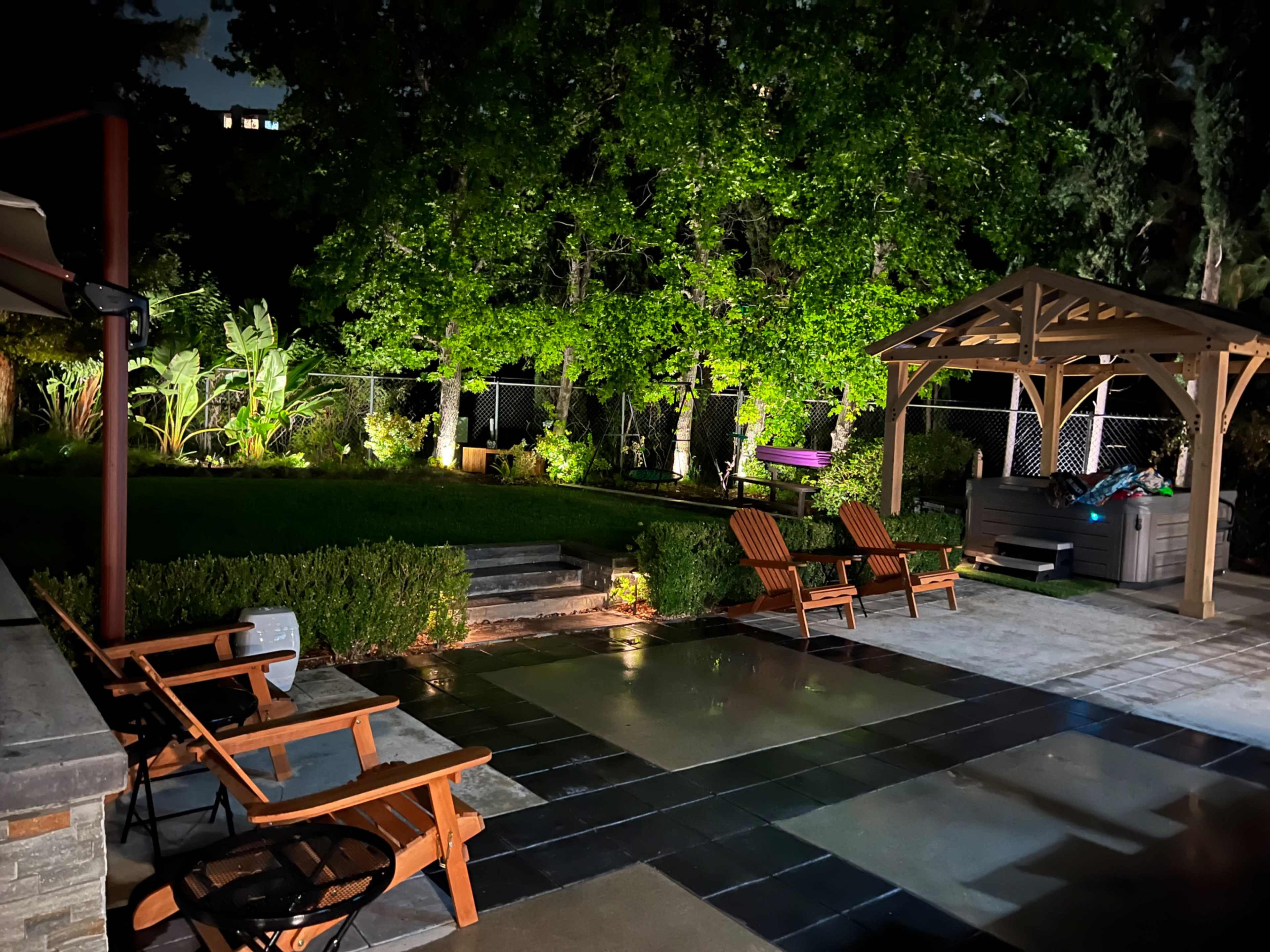 The image shows a dimly lit backyard featuring a landscaped area with a gazebo and two wooden chairs on a stone patio.